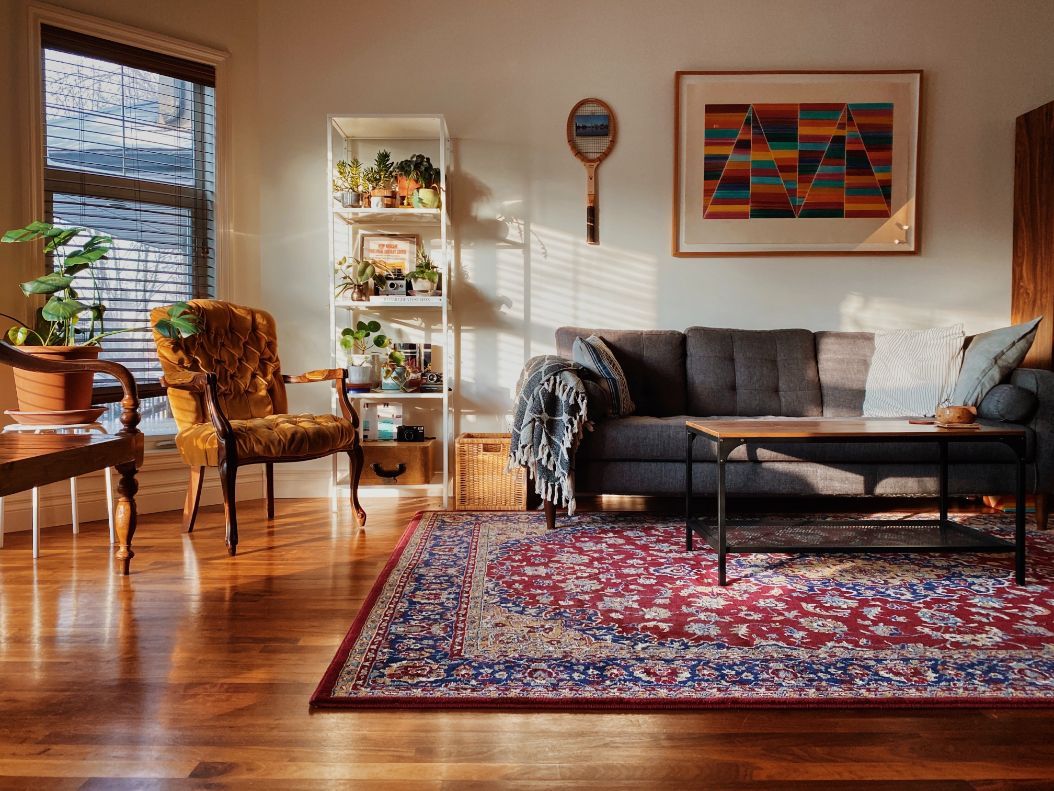 Living room bathed in sunlight with a sofa, antique rug, armchair, plants, and artwork on the wall.