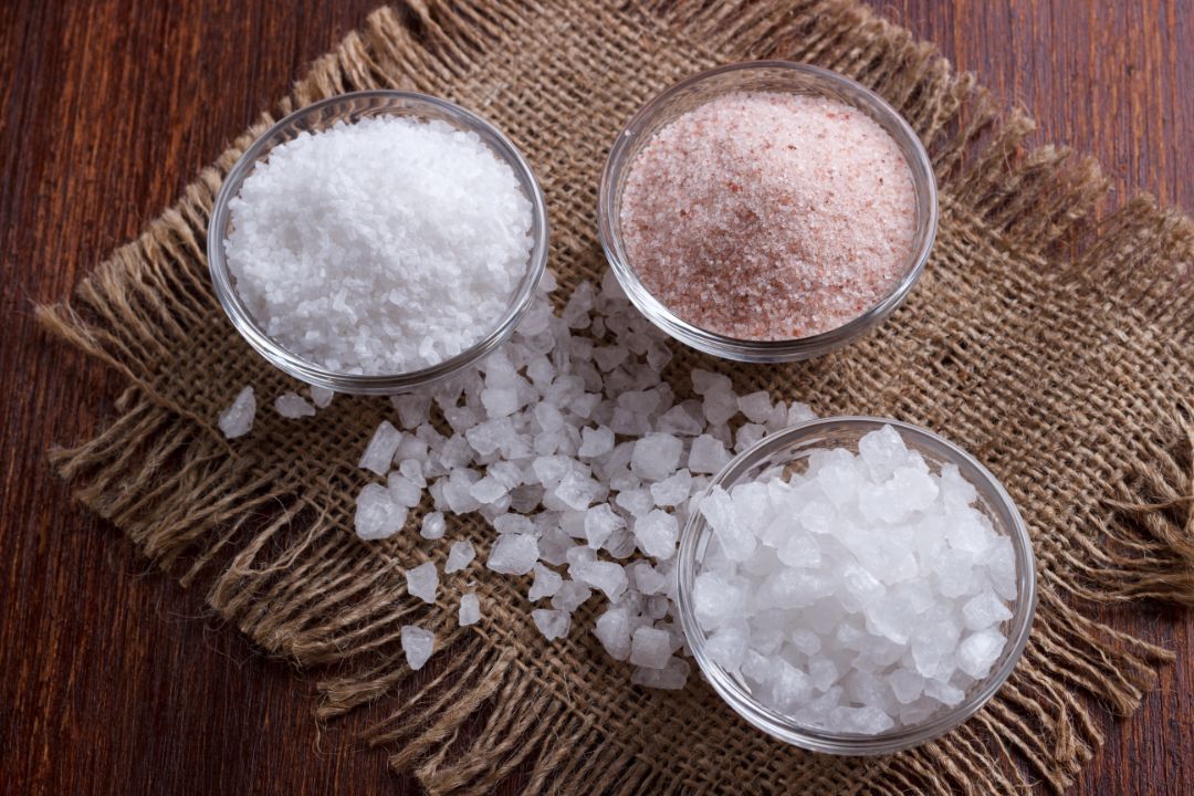 Three bowls of different salts: white, pink, and large white crystals on a burlap cloth, wooden surface.