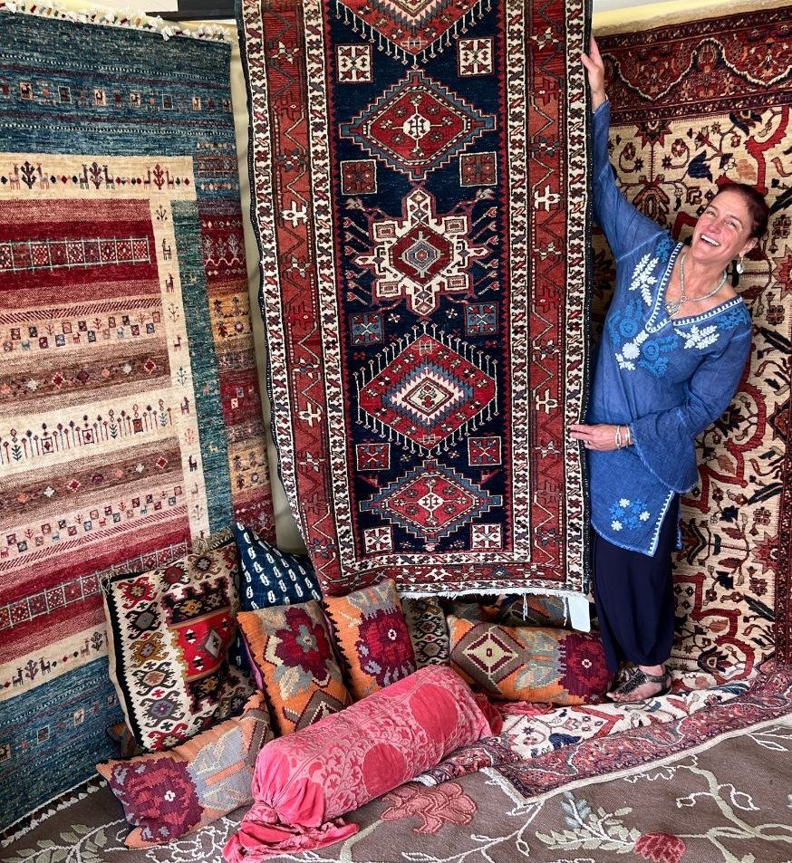 Cynthia First behind hanging patterned rugs, with colorful pillows on the floor at Mulford Farmstead. 