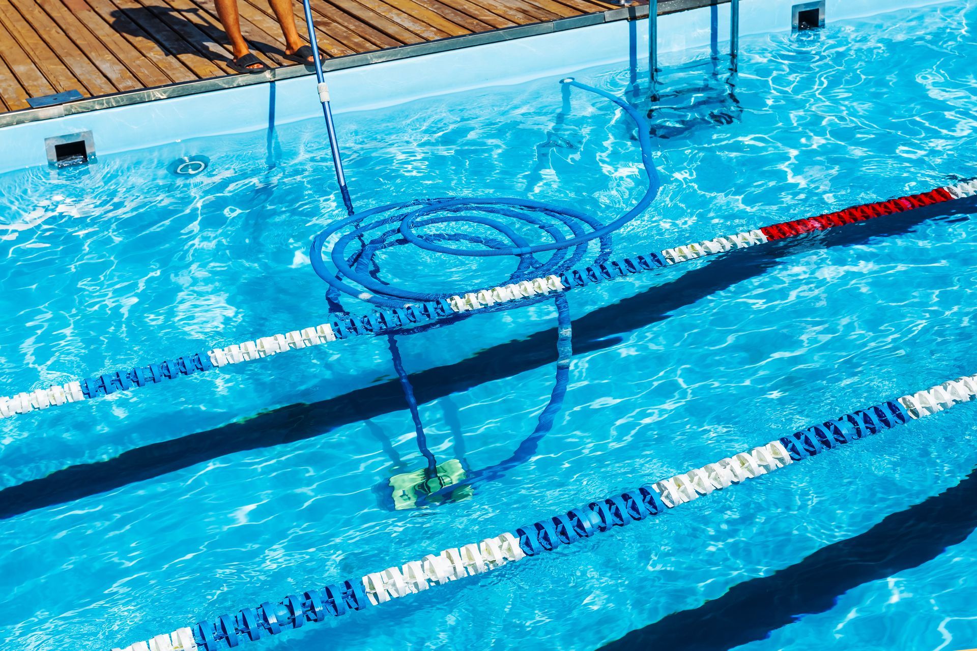 Pool cleaner in blue water, hose coiled near pool edge, person standing on the deck.