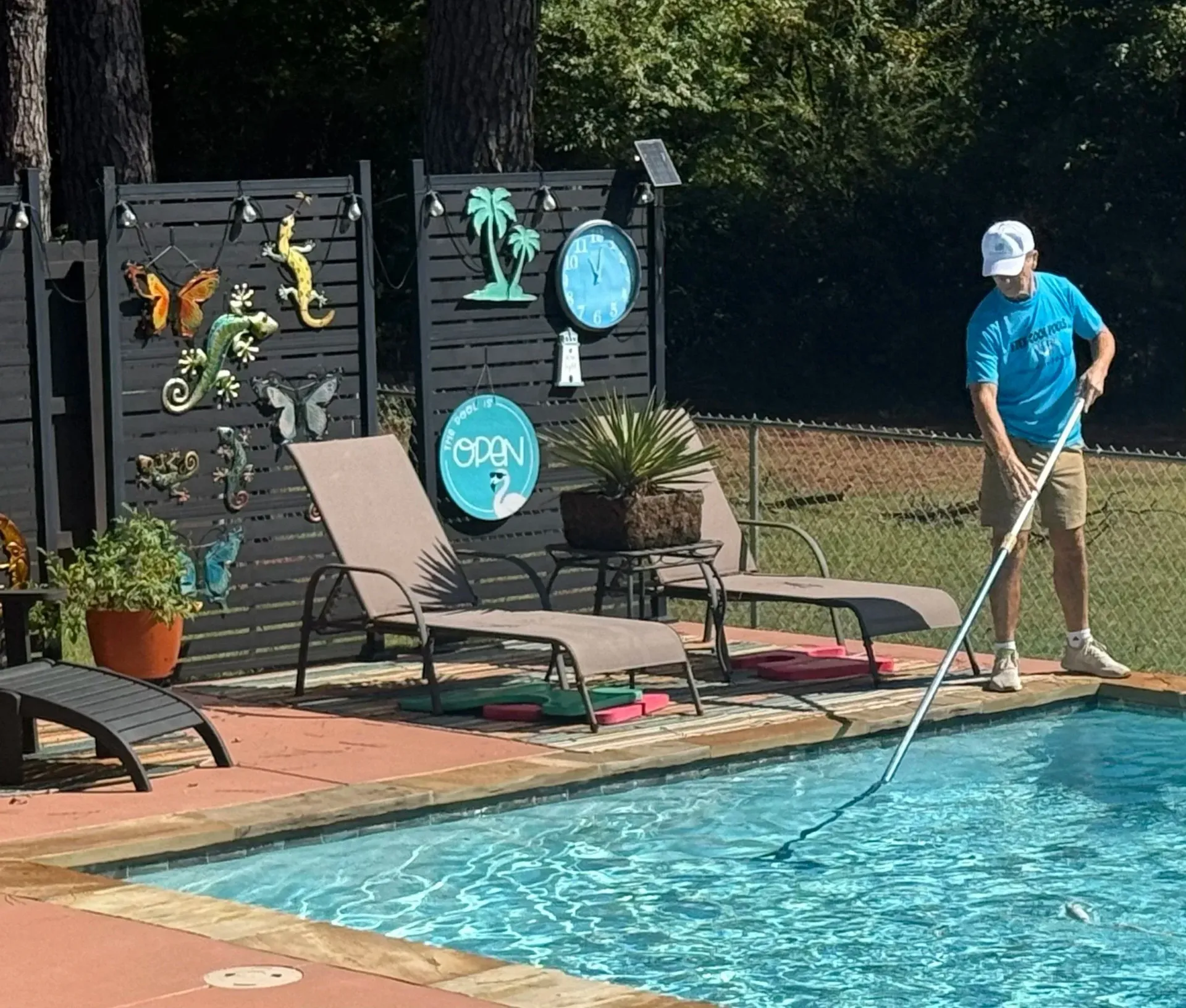 Man cleaning a swimming pool with a net; lounge chairs and decorative fence in the background.
