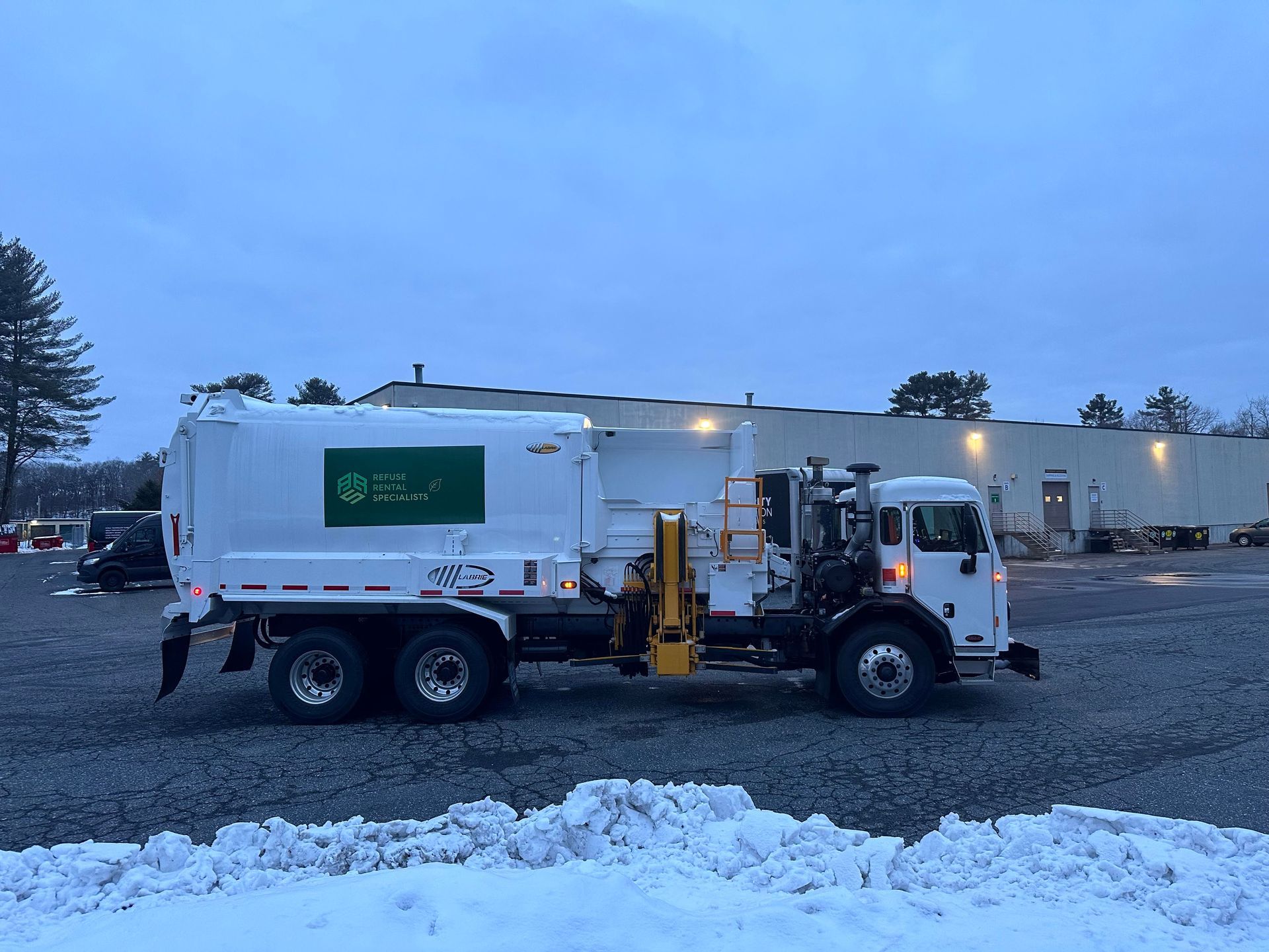 A white garbage truck is parked in a snowy parking lot.
