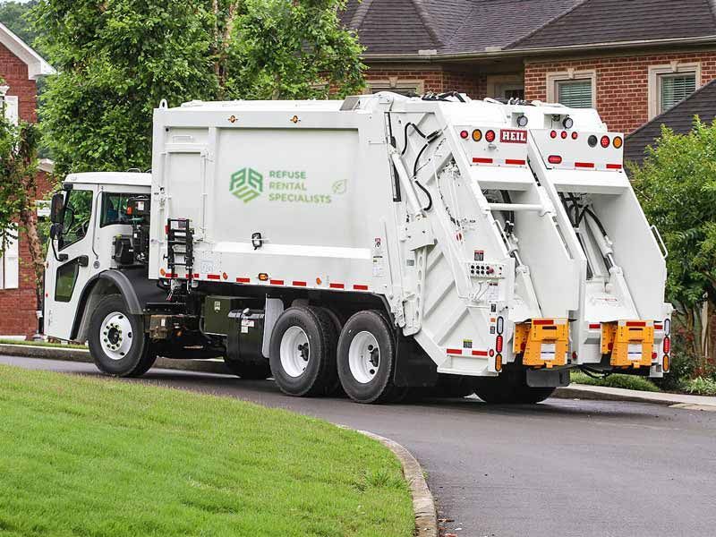 A garbage truck is driving down a street next to a brick house.