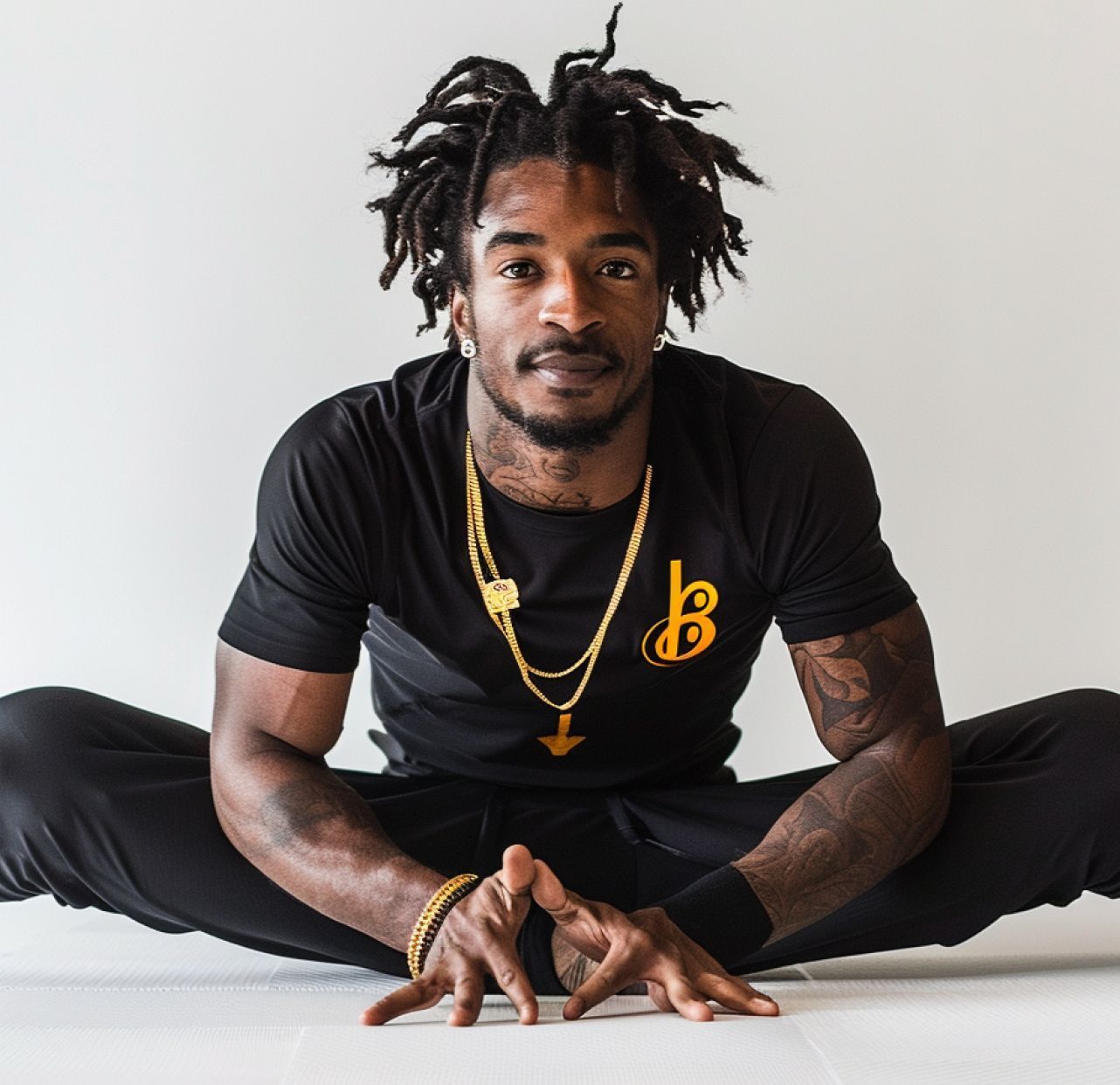 Man seated on the floor in a wide-legged stretch, wearing a black outfit and gold chains, against a white background