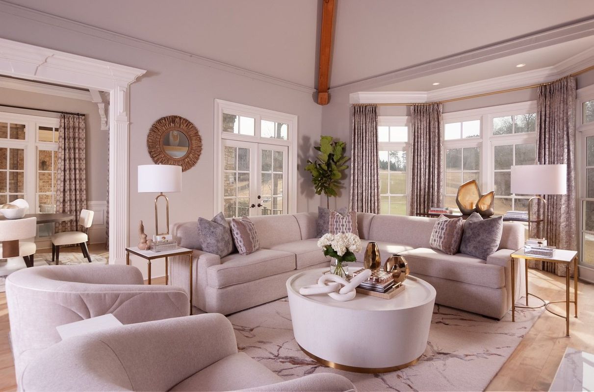 Bright, elegant living room with beige sofas, round white coffee table, lamps, and large windows with sheer curtains