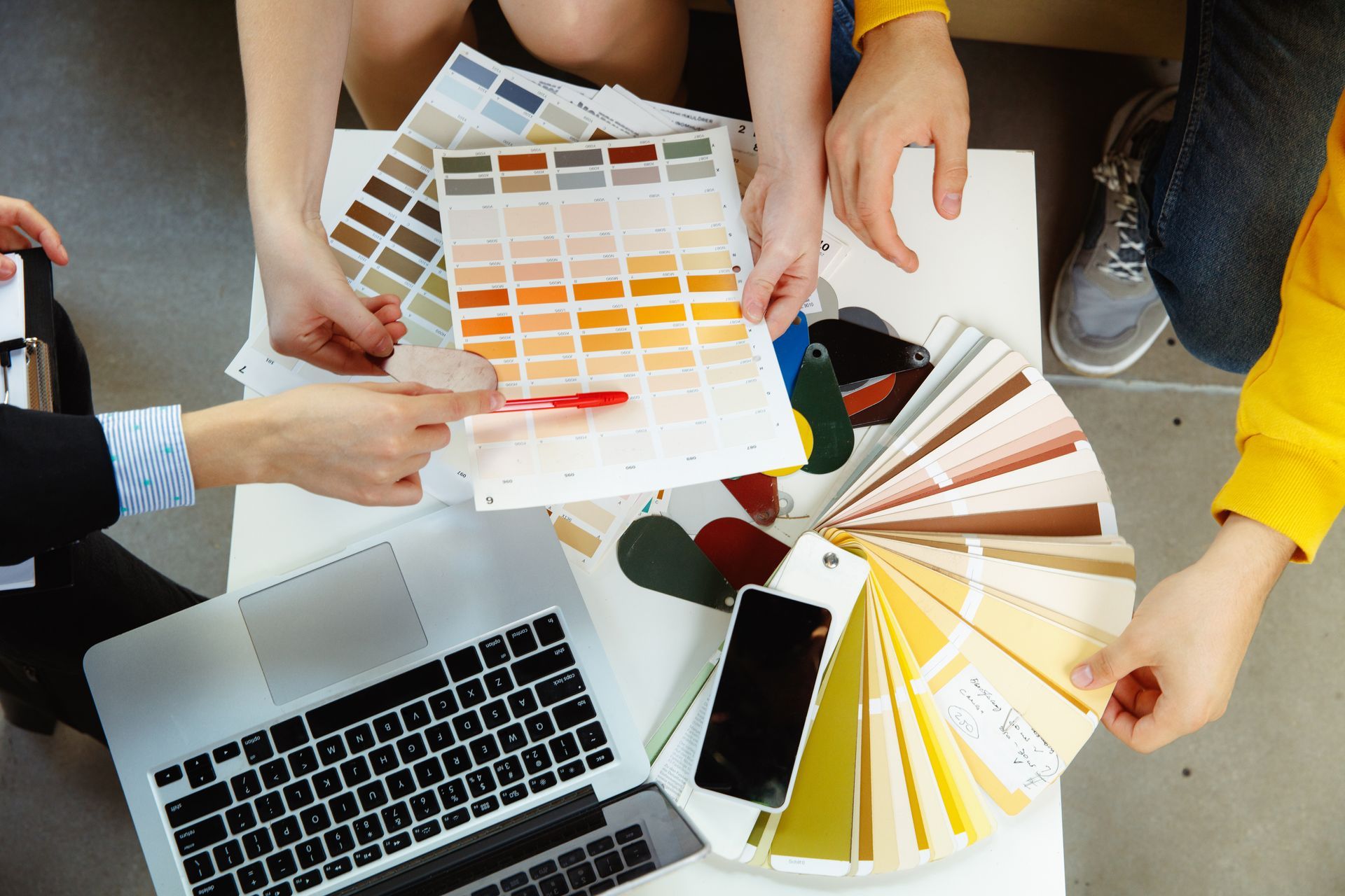 Hands sampling paint swatches around a laptop, phone, and color fan deck on a table