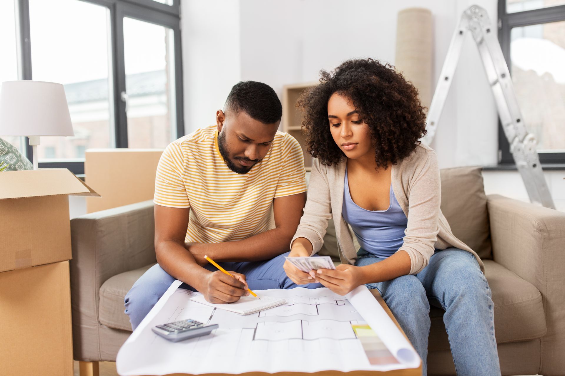 Two people reviewing paperwork and calculator on a table in a bright living room with moving boxes