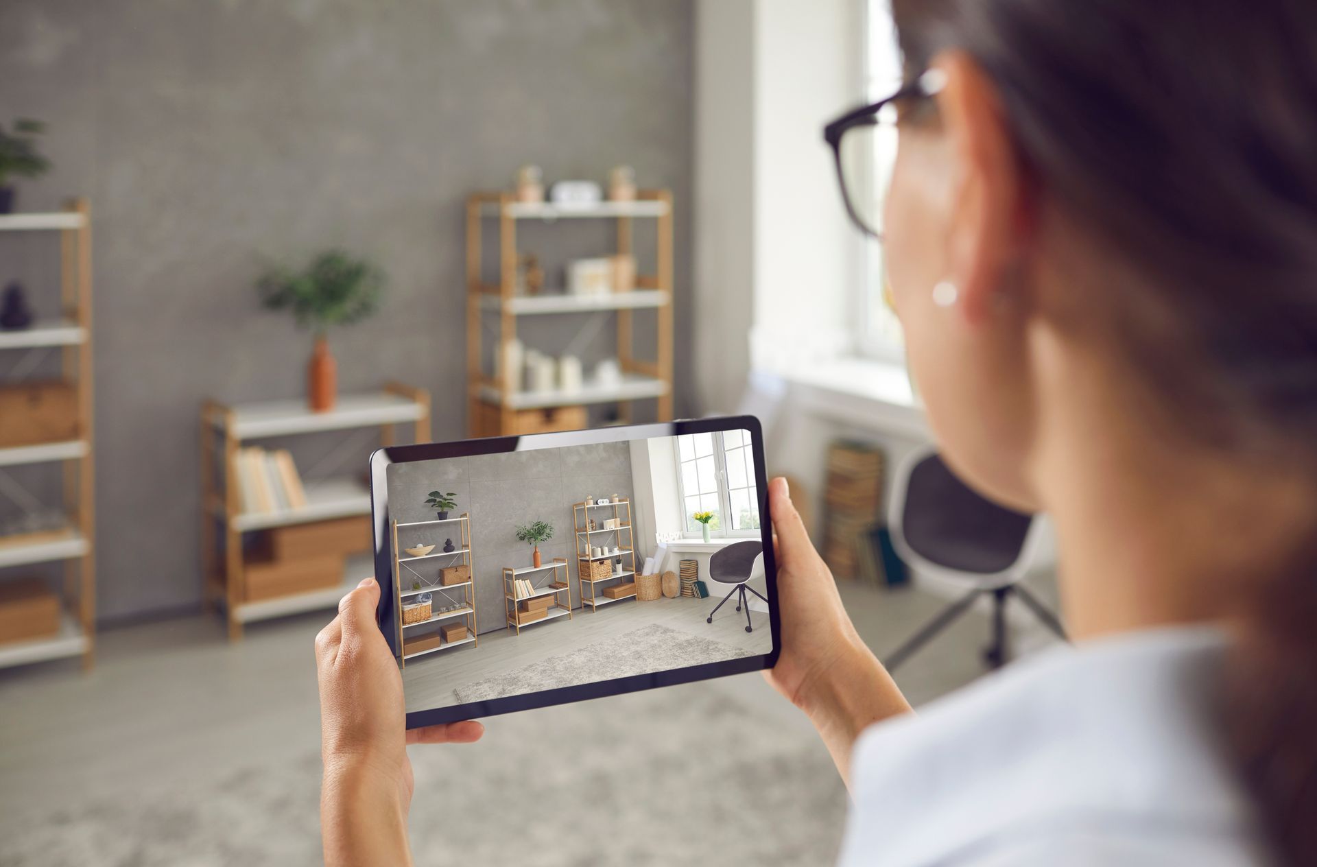 Person holding a tablet that shows a room design, viewed from behind in a bright home office