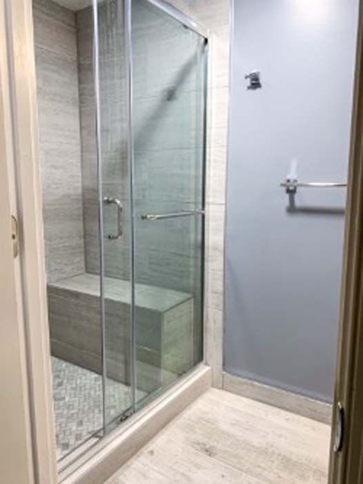 Modern bathroom with a glass shower door, gray tiled walls, and a built-in bench. Light gray and white flooring, blue wall outside the shower.