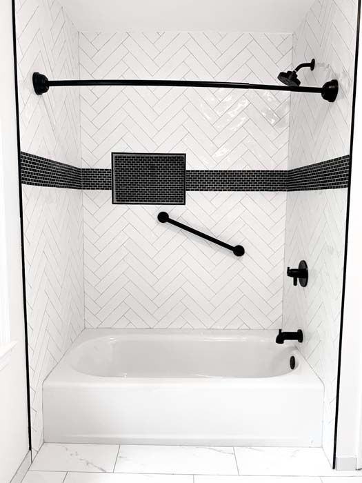 A white bathtub and shower with black accents, including a grab bar, showerhead, and towel rod. The walls feature white herringbone tile with a black tile border.