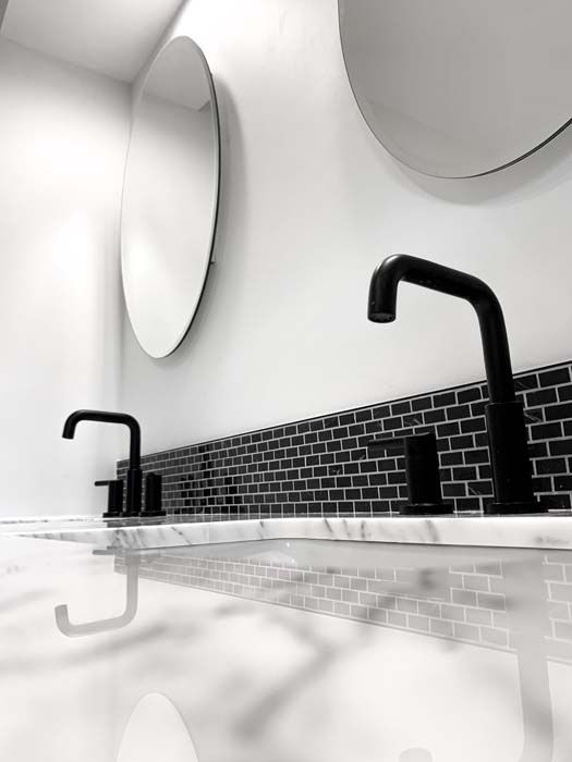 Black and white bathroom with two black faucets and round mirrors. The counter has black brick tile and a marble-like finish.