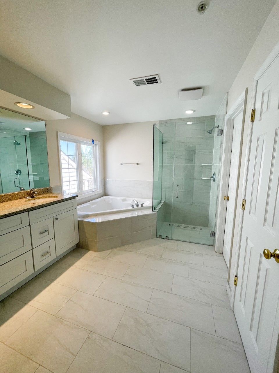A modern bathroom with a white vanity, shower, and corner jacuzzi. Light-colored tile and white walls create a bright space.
