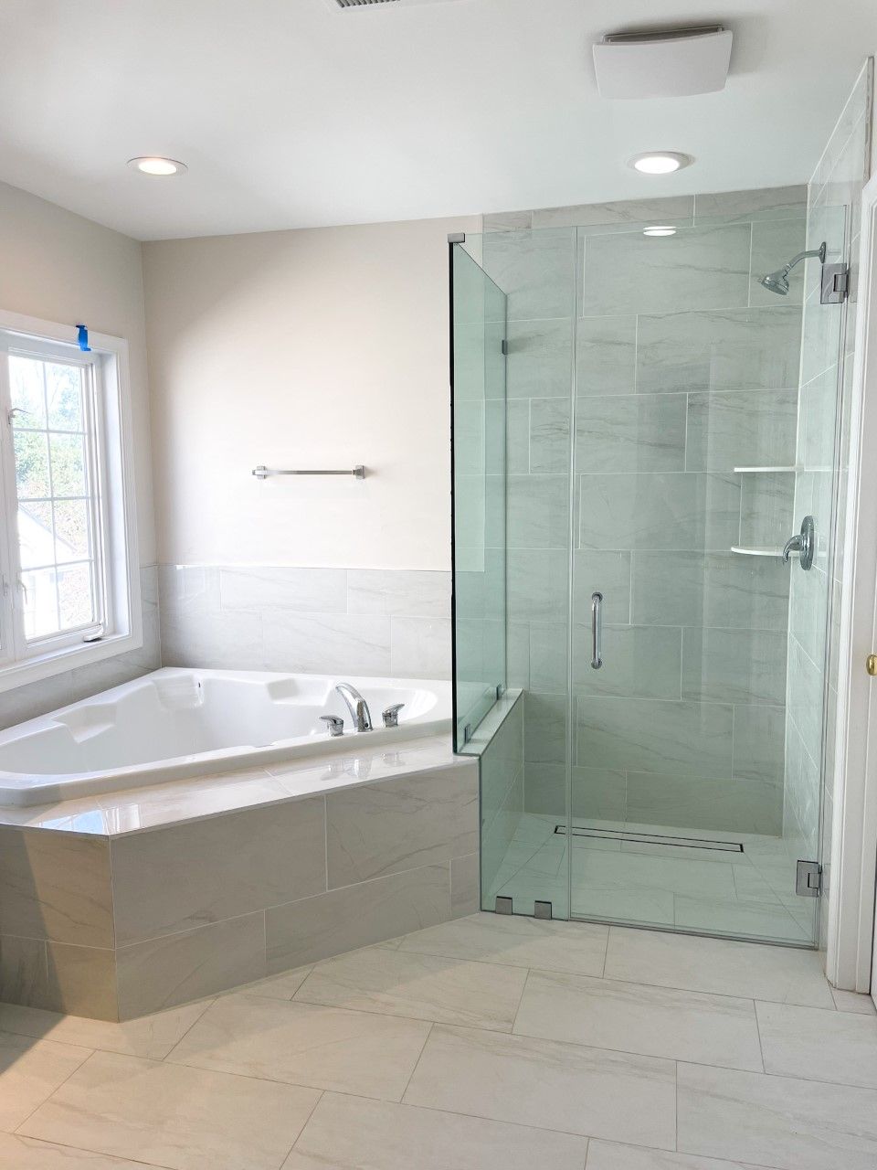 Modern bathroom with a corner bathtub and a glass-walled shower. The room features light-colored tiles, neutral walls, and a window.