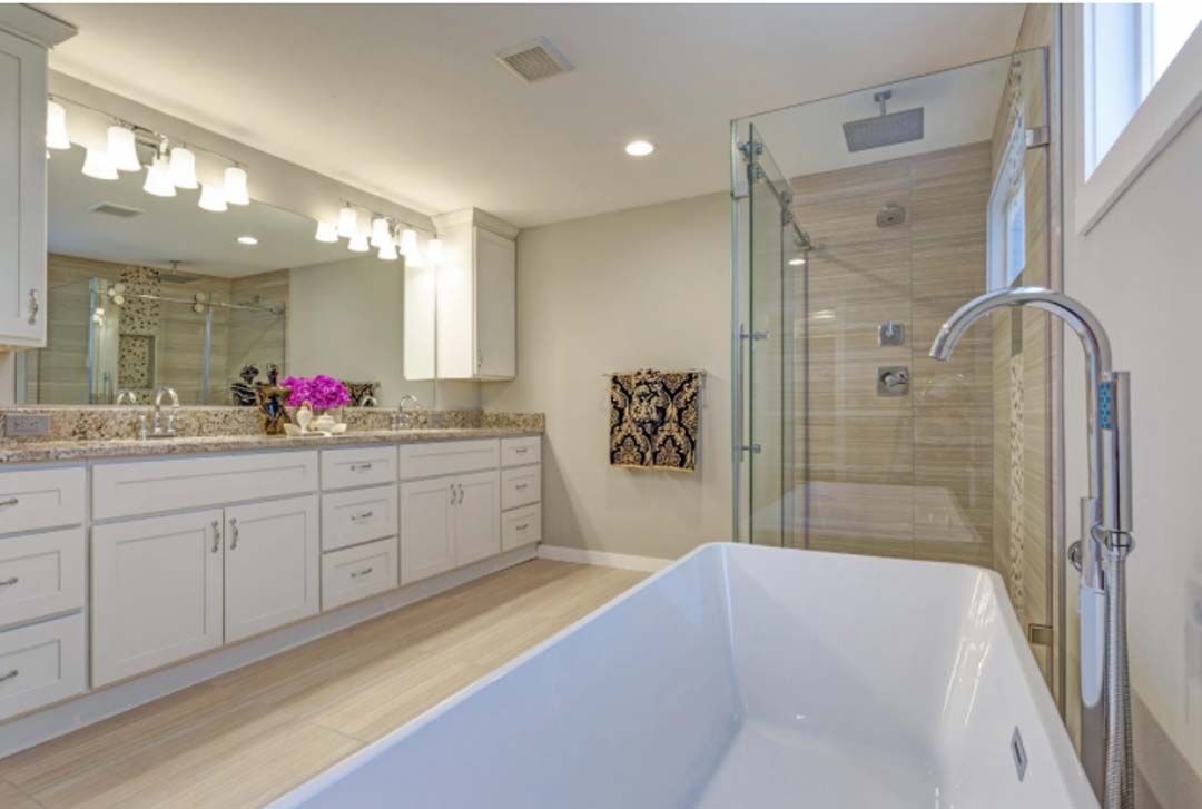 Bright bathroom with white cabinets, a freestanding tub, and a glass shower. Beige walls and light-colored flooring.