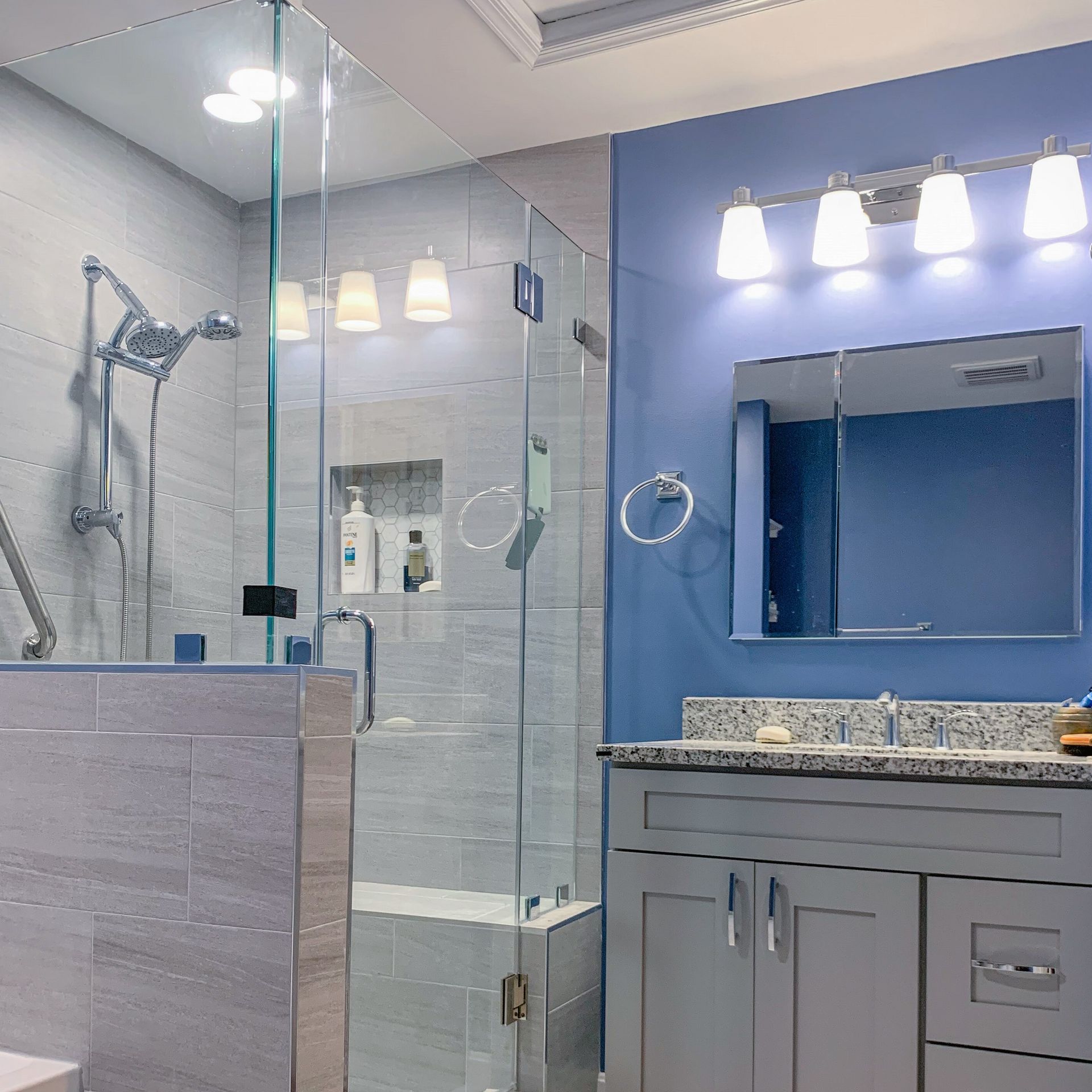 Modern bathroom with a glass shower, gray cabinets, and blue walls. Shower fixtures and vanity lights are visible.