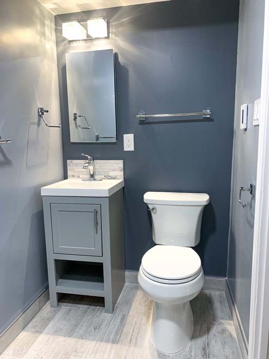 Small bathroom with gray walls, white toilet, gray vanity, and light-colored wood-look flooring. A silver towel bar and mirror are visible.