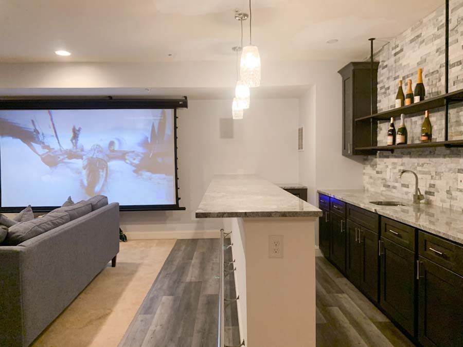 Basement bar with a granite countertop and a movie screen. Dark cabinets, grey wood-look flooring, and white walls.