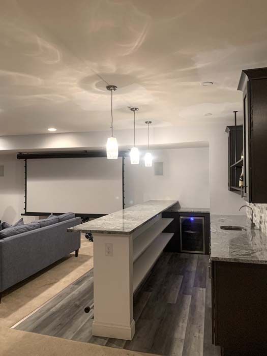 A finished basement with a bar and entertainment area. Features include a bar with a granite countertop, shelves, a wine fridge, a projector screen, and pendant lights.