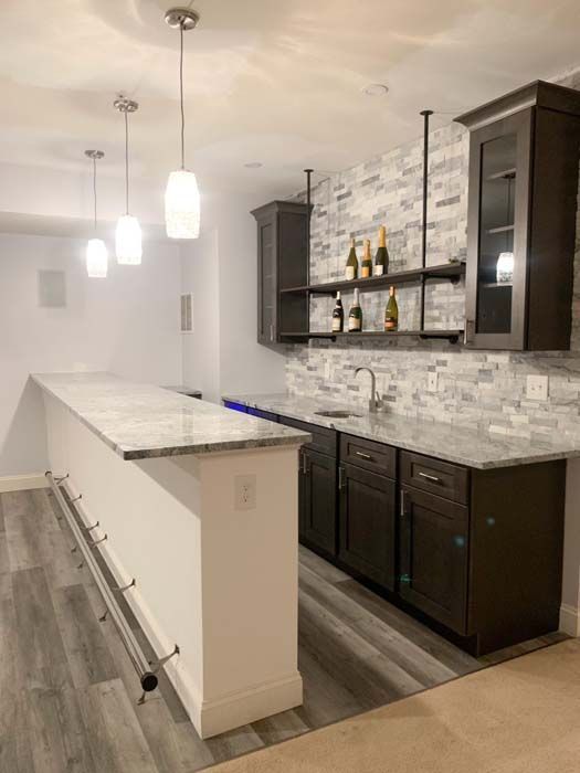 Basement bar with white and dark brown cabinets, marble countertops, and gray plank flooring. Three pendant lights hang above the bar.