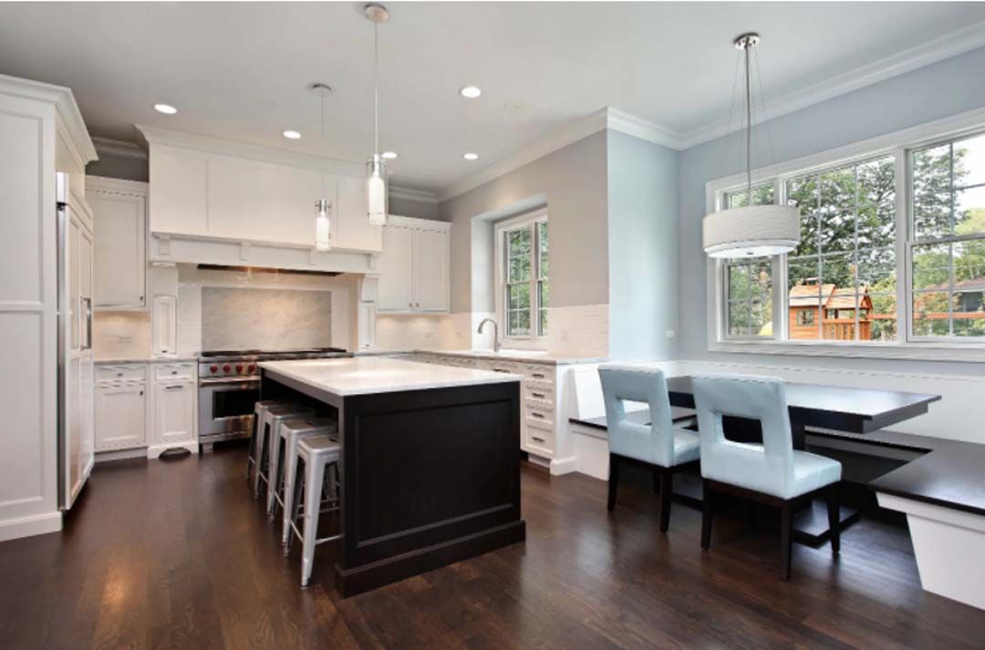A modern kitchen with white cabinets, a black island, and a breakfast nook with light blue chairs. Large window offers outside view.
