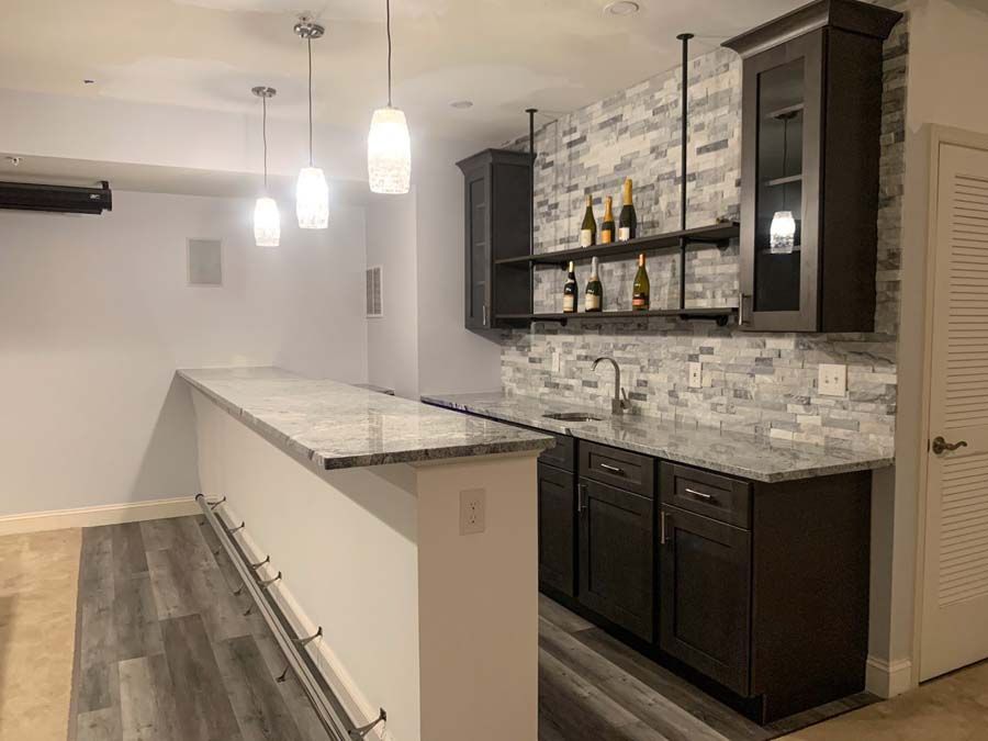 Basement bar with granite countertops, dark cabinets, and a gray and white stone backsplash. Three pendant lights hang over the bar.