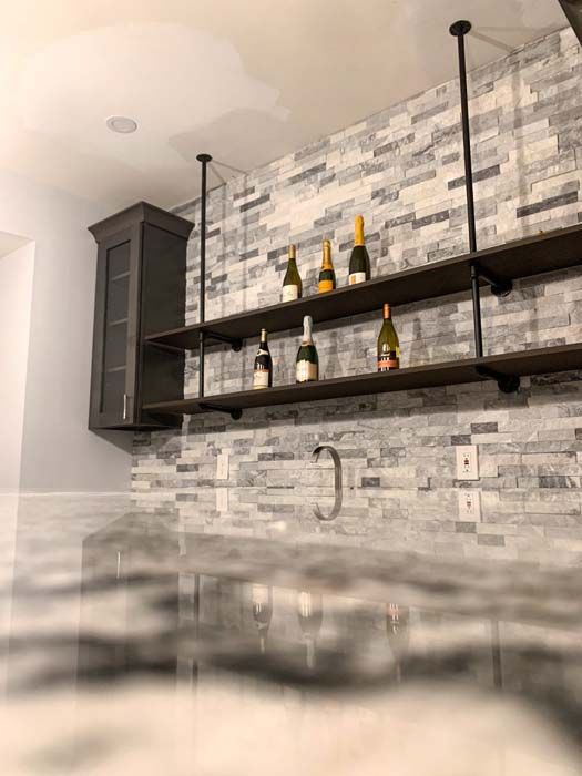Bar area with gray and white tile backsplash, dark shelves supported by black pipes, and bottles of champagne. Gray cabinet on the left, marble countertop in foreground.