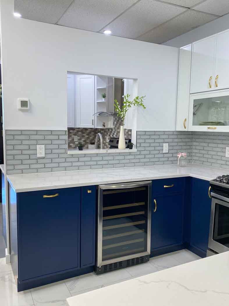 Navy blue and white kitchen with a wine fridge, light-colored countertops, and a pass-through window with a vase of green plants.