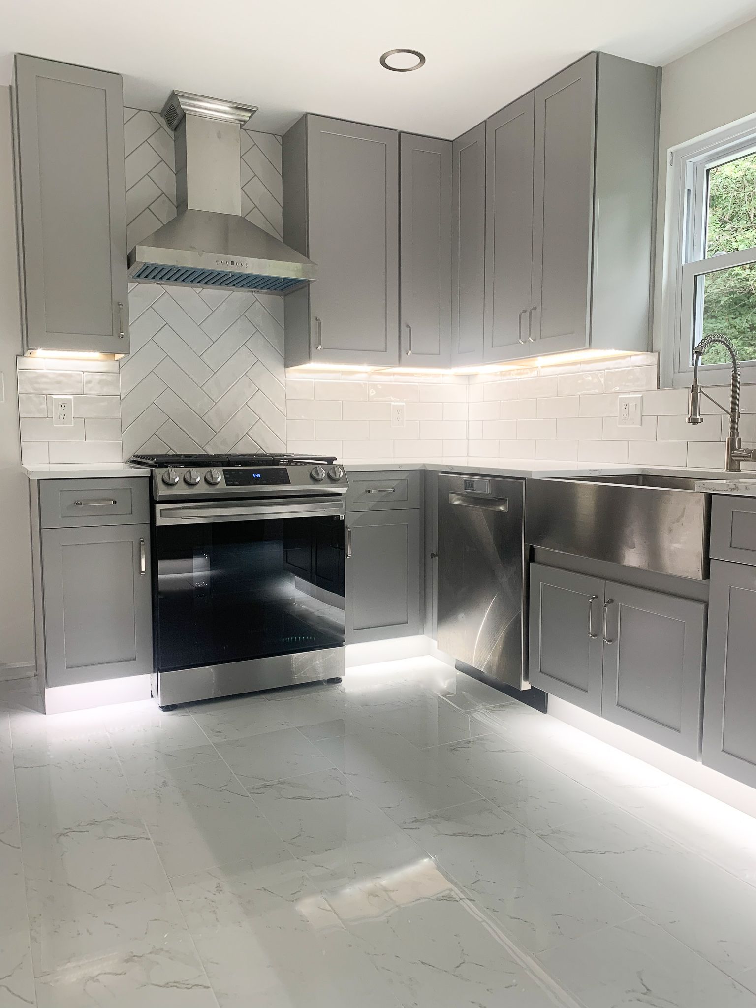Modern gray kitchen with stainless steel appliances, herringbone backsplash, and under-cabinet lighting.