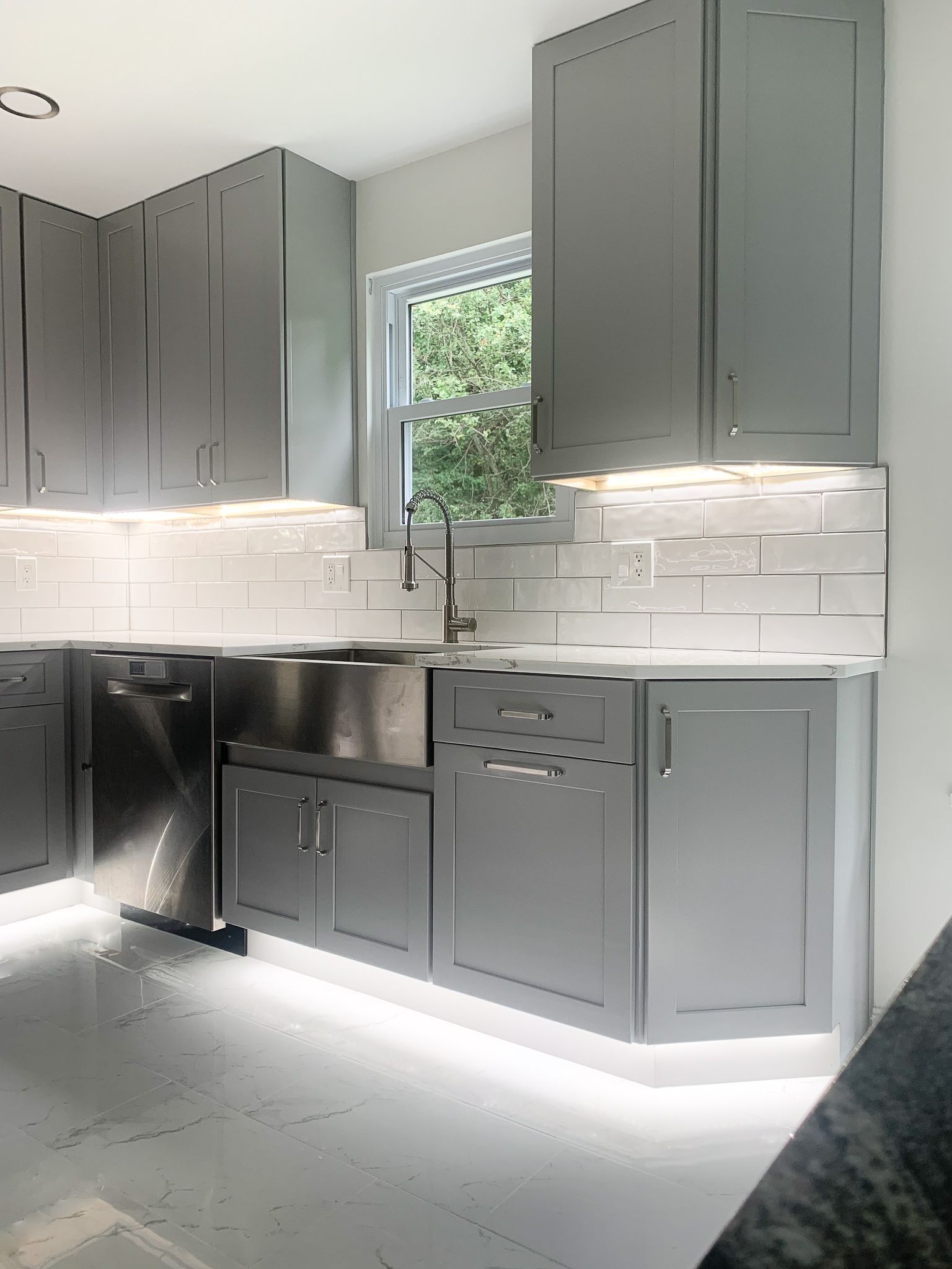 Modern gray kitchen with stainless steel appliances, white backsplash, and under-cabinet lighting. A window shows a view of green trees.