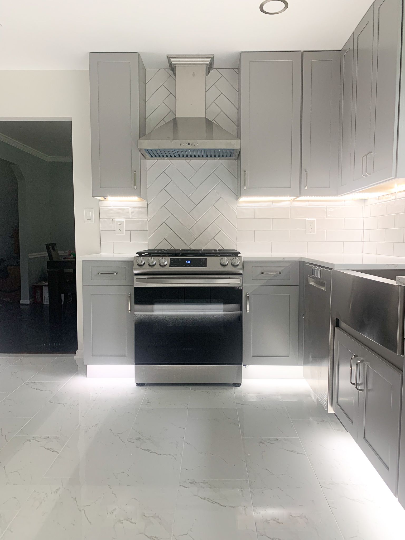 Modern kitchen with gray cabinets, stainless steel appliances, and white backsplash.  White tiled floors reflect the bright lighting.