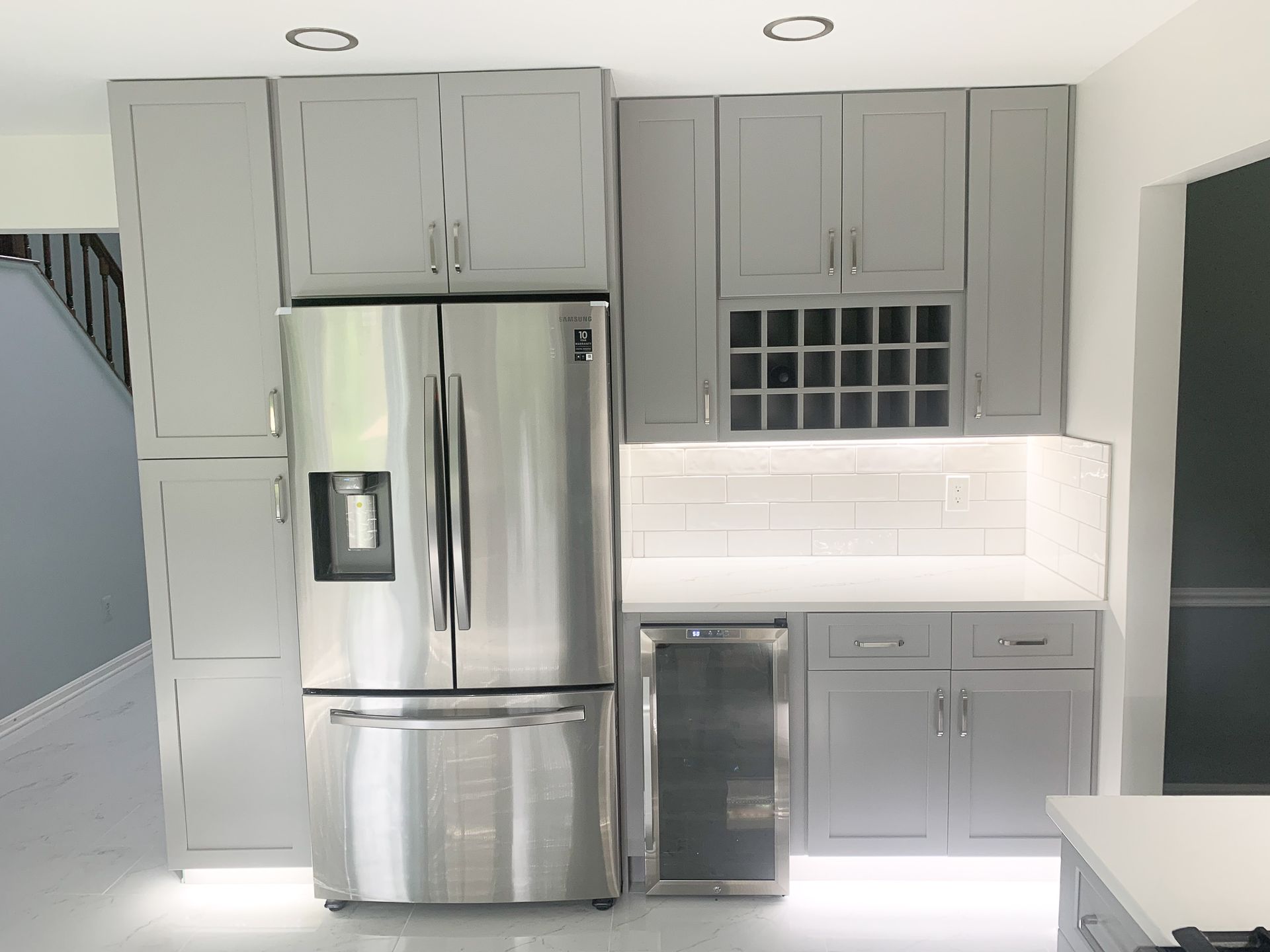 Gray kitchen cabinetry surrounds a stainless steel refrigerator, wine cooler, and overhead wine rack, illuminated by under-cabinet lighting.