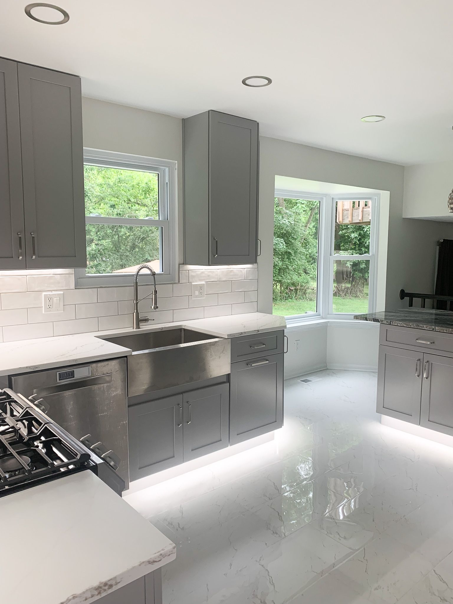 Modern gray kitchen with stainless steel appliances, white countertops, and under-cabinet lighting. Includes a stainless steel farmhouse sink and a window with a view of greenery.