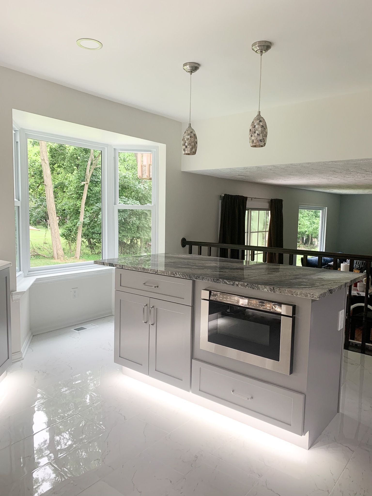 Modern kitchen with gray island, built-in microwave, and pendant lights. Large window offers outdoor view; white floor and cabinets.