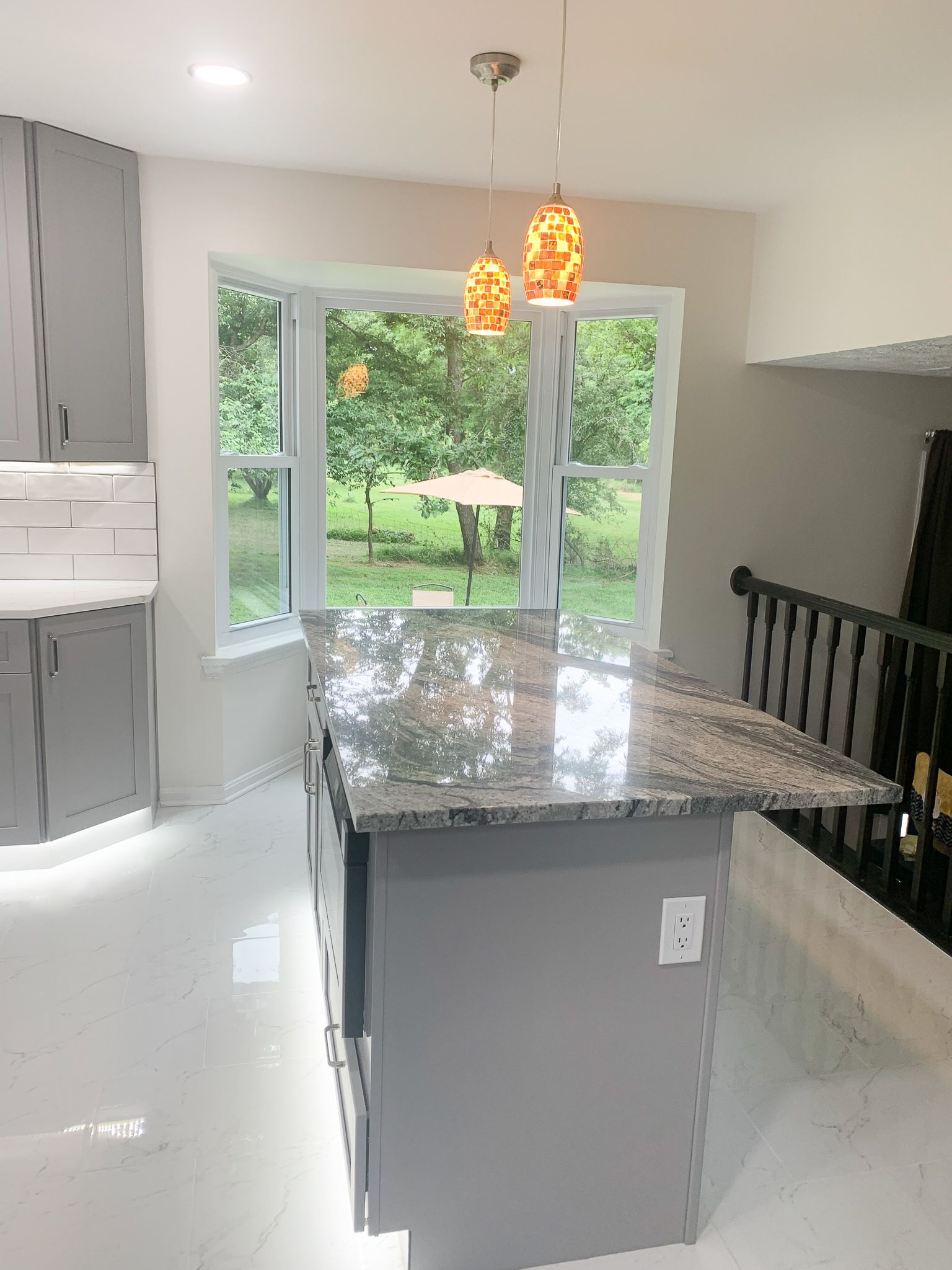 A modern kitchen with a gray island and granite countertop, featuring two pendant lights, in front of a bay window with a backyard view.