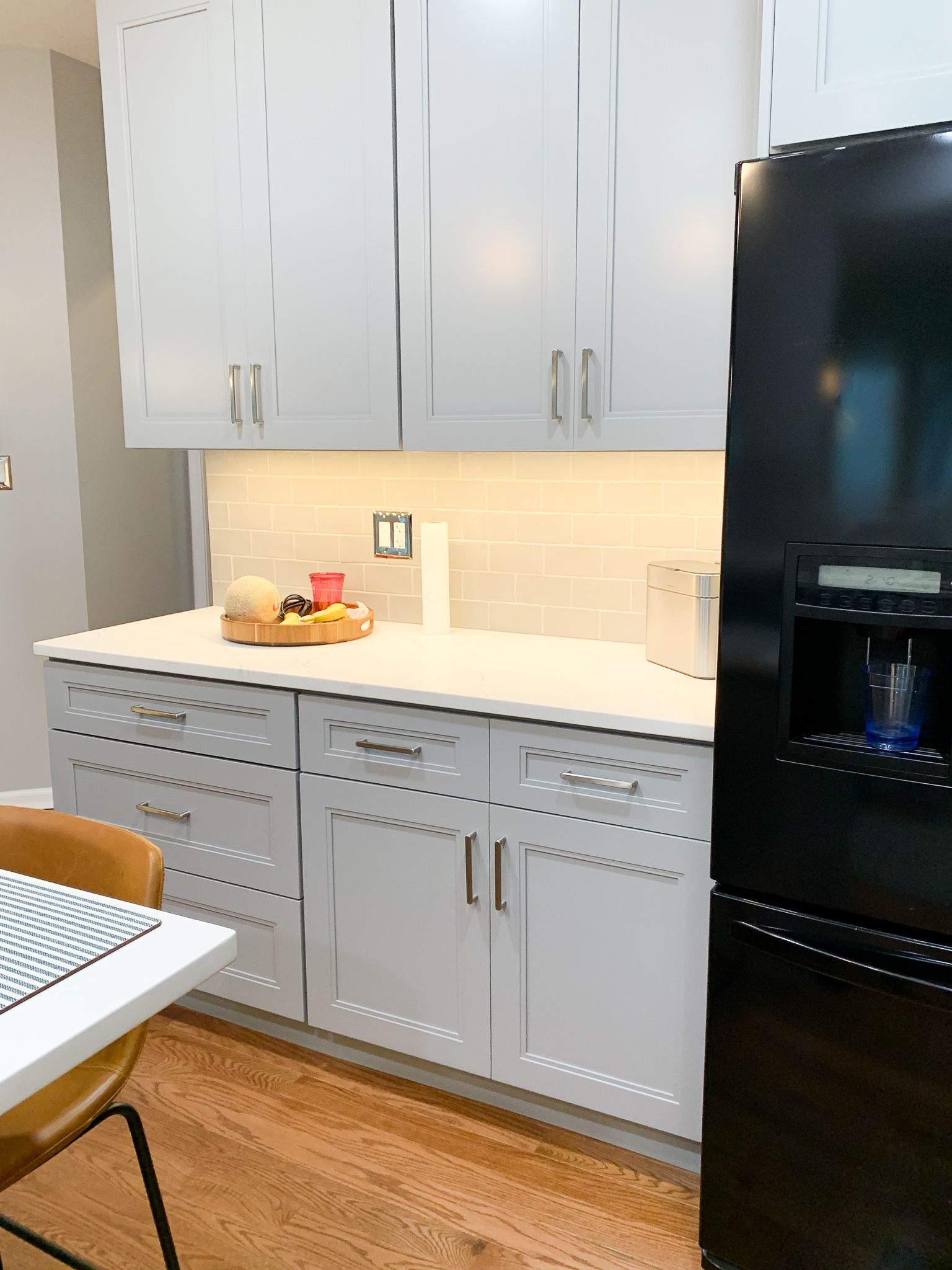 White kitchen cabinets with a light countertop, illuminated backsplash, and a black refrigerator on the right.