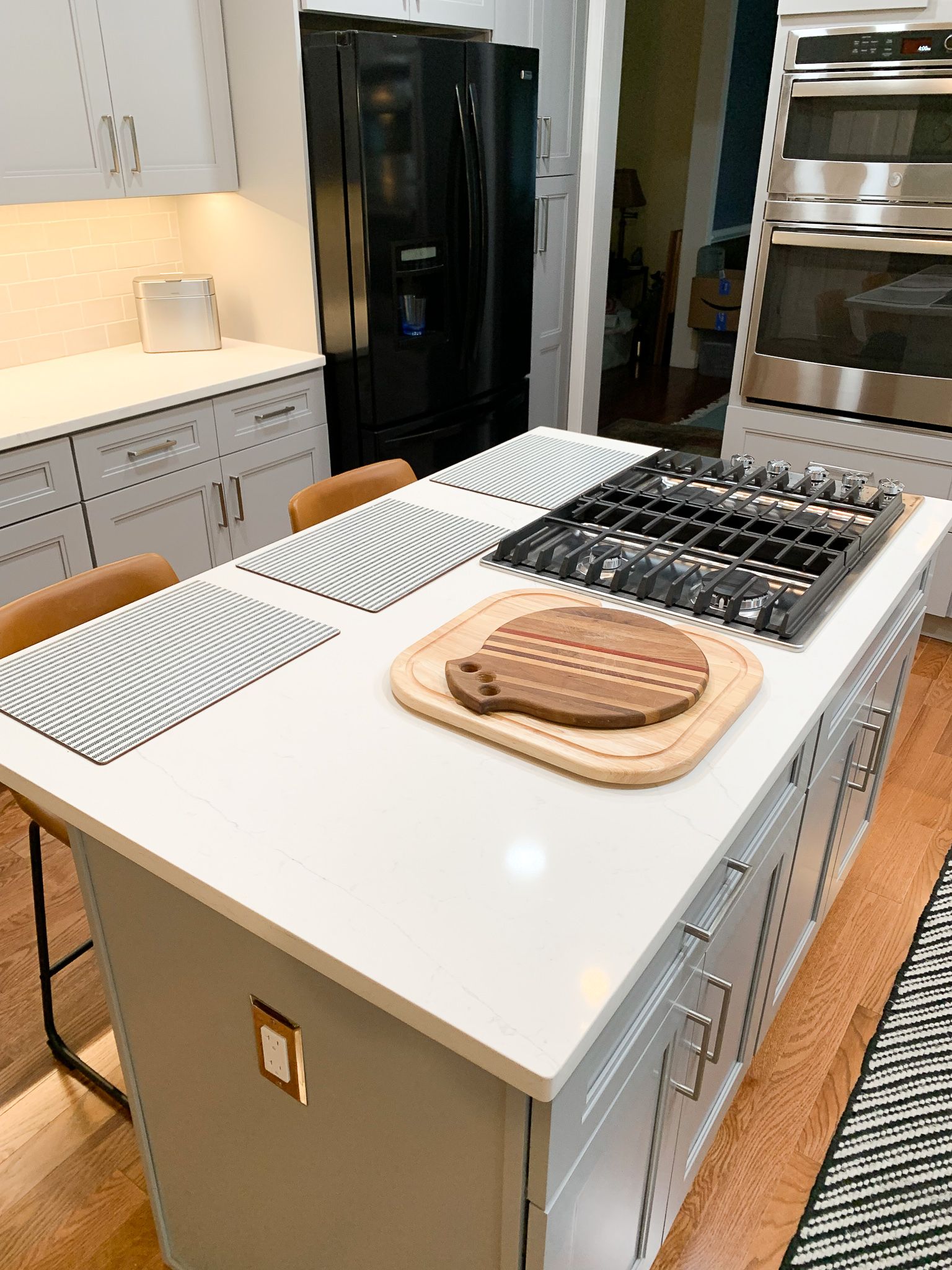 Modern kitchen with a white island with a cooktop, a cutting board, and seating; a black fridge and built-in ovens are visible.
