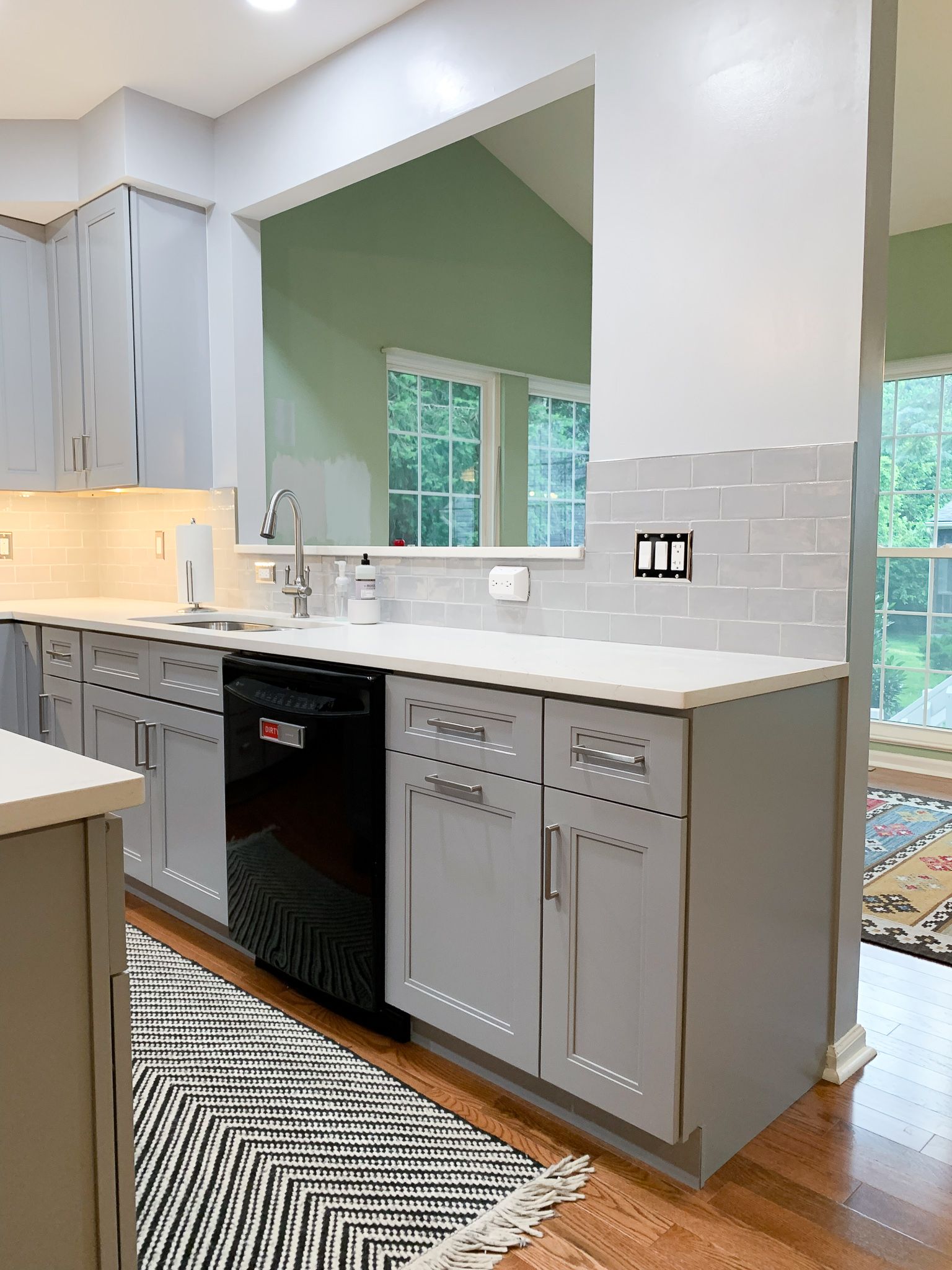 Gray kitchen with white countertops, black dishwasher, and backsplash. A window reveals greenery.