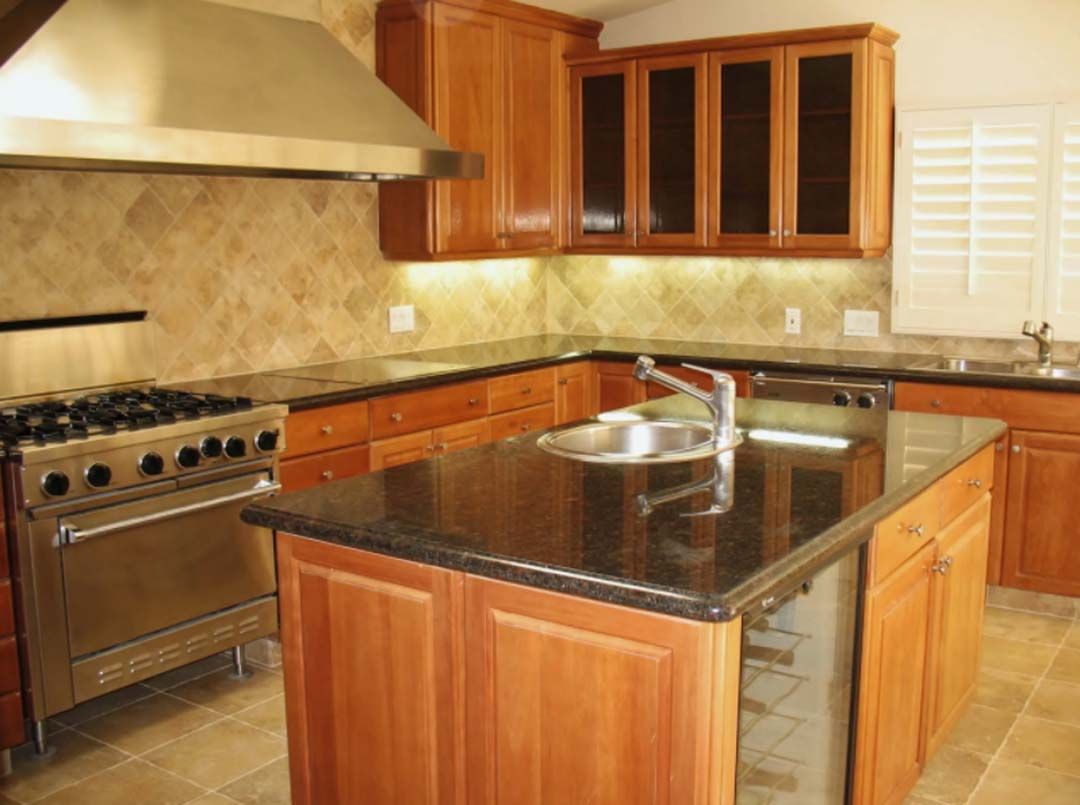 A kitchen with wood cabinets, dark countertops, and stainless steel appliances. An island with a sink and wine fridge sits in the center.