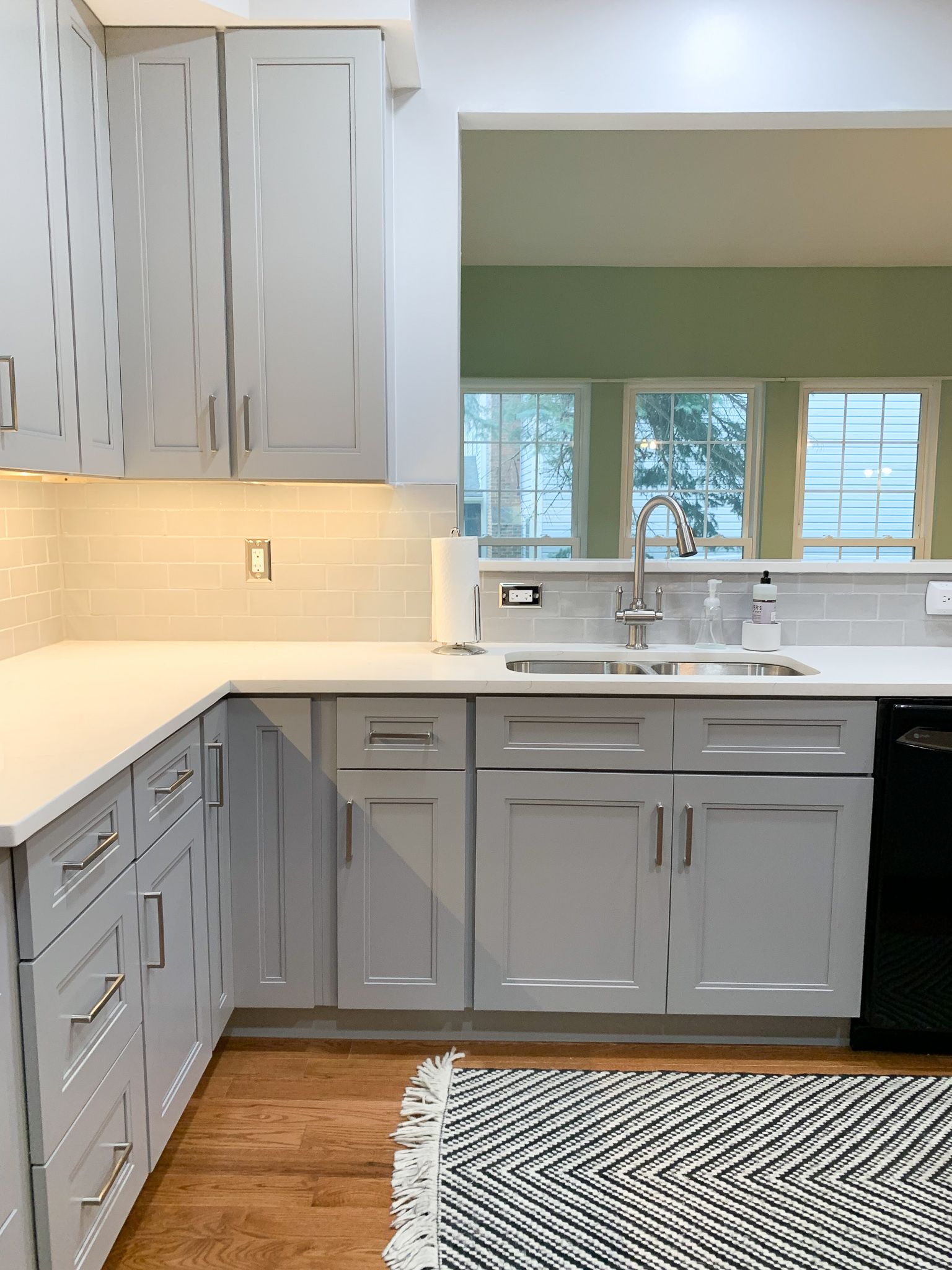 Light gray kitchen with white countertops, cabinets, and a black and white rug. A sink faces a bright green-walled space with windows.