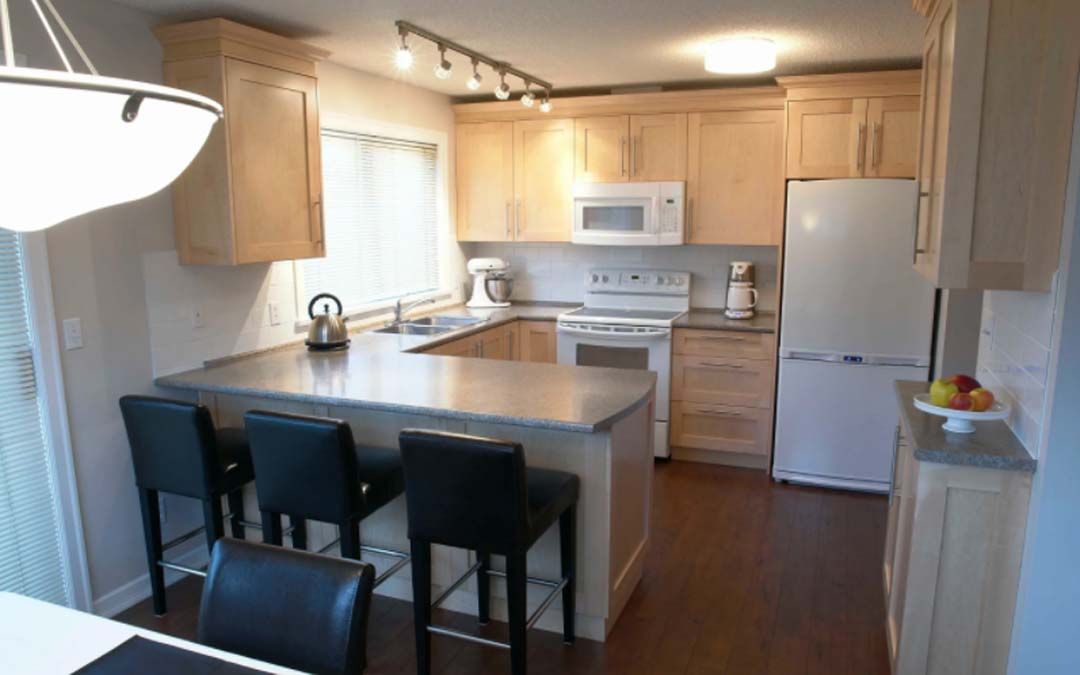 U-shaped kitchen with light wood cabinets, gray countertops, and a breakfast bar with black stools. A white refrigerator stands to the right.
