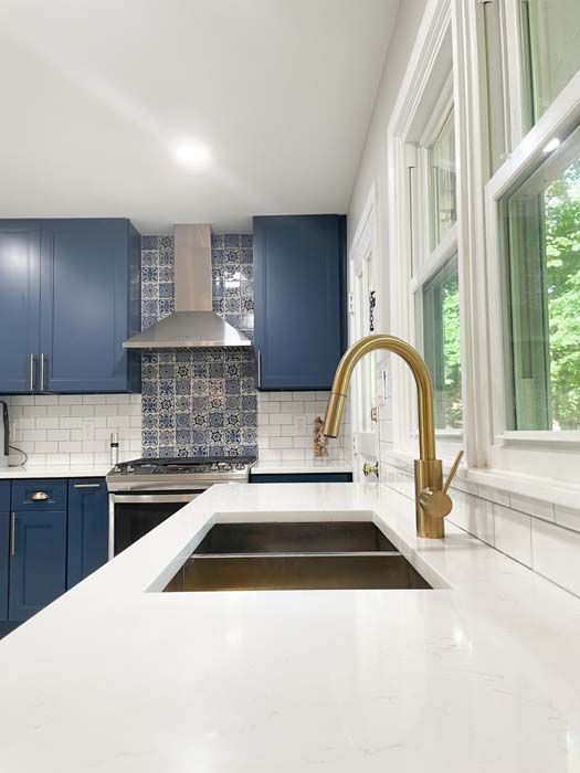 Kitchen with navy blue cabinets, white countertops, and a brass faucet. Features a stainless steel range hood and tile backsplash.
