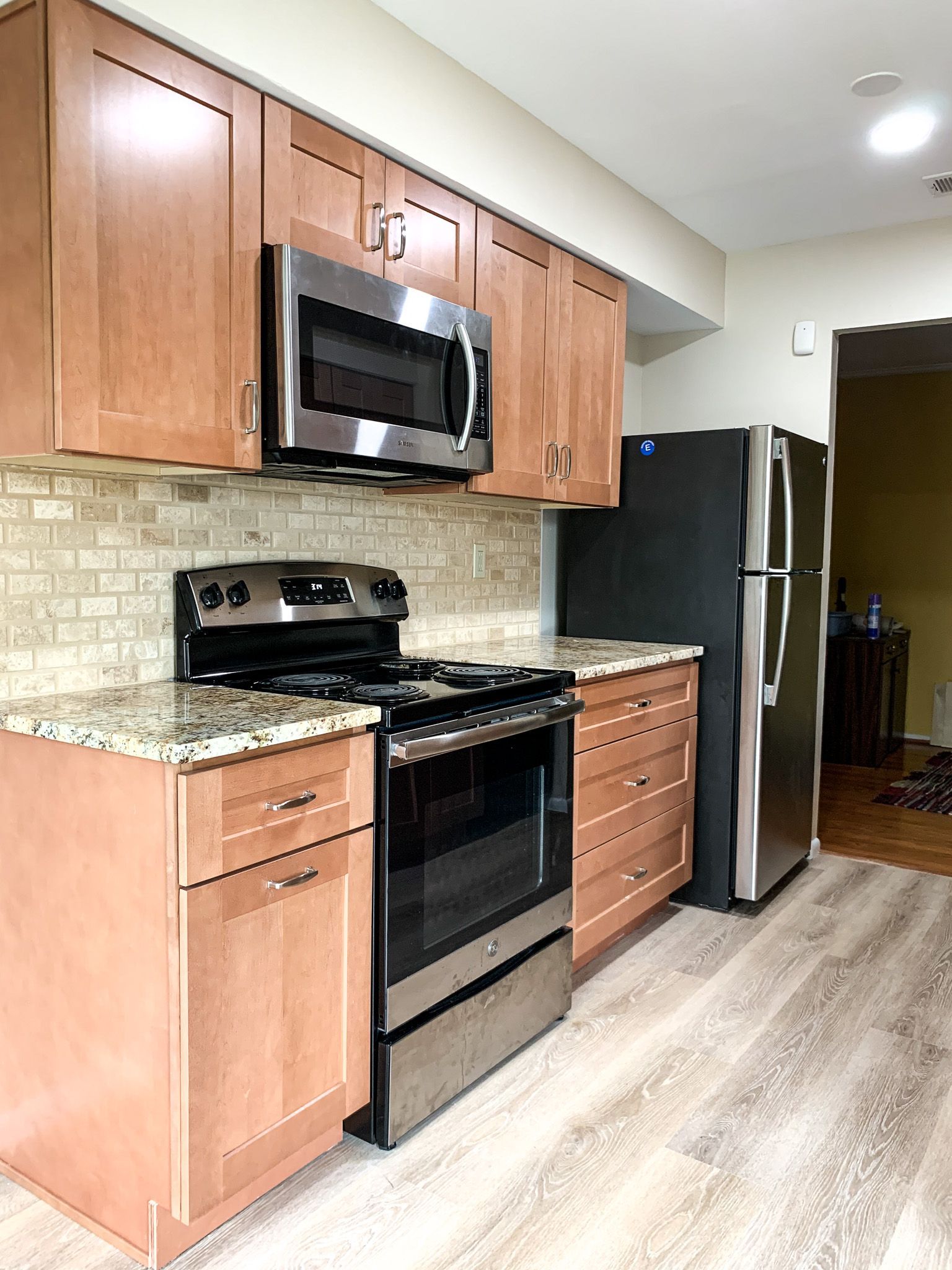 A modern kitchen with light wood cabinets, stainless steel appliances, and a tiled backsplash. The floor is a light gray laminate.