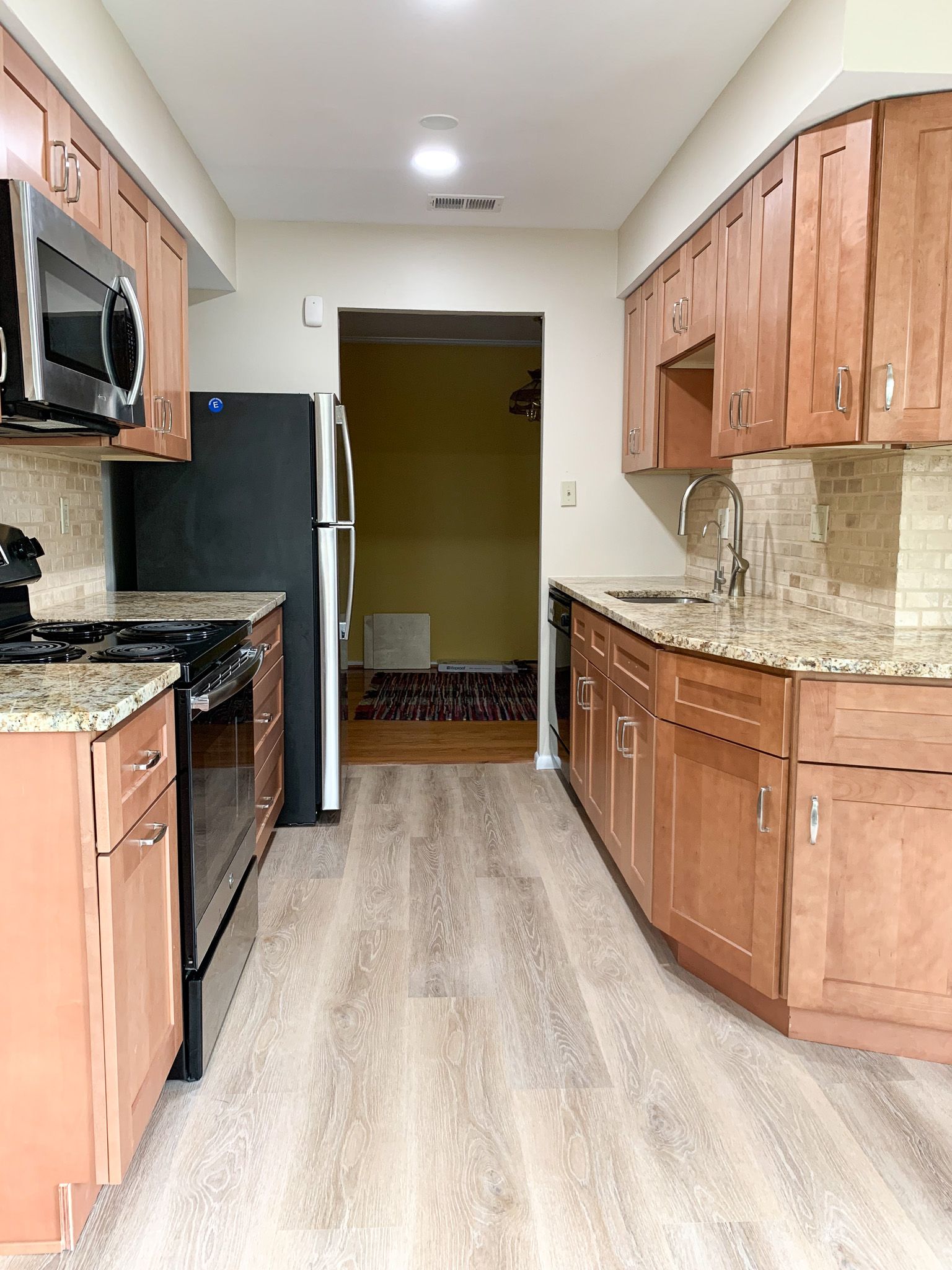 A galley kitchen with brown cabinets, granite countertops, and stainless steel appliances. The floor is light wood, and a doorway is visible in the distance.