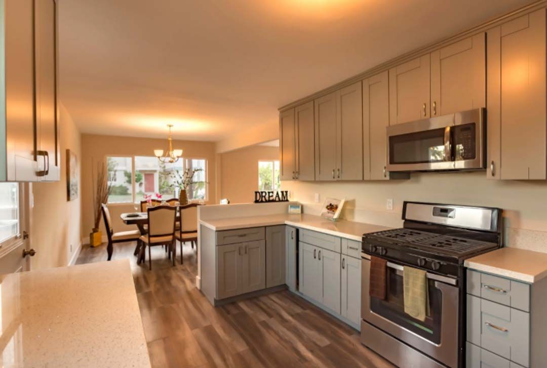 A modern kitchen with gray cabinets, stainless steel appliances, and wood-look flooring. A dining room is visible in the background.