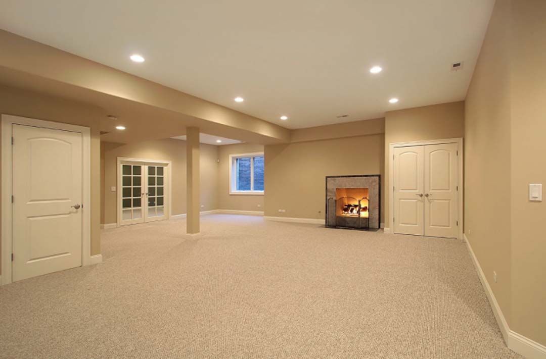 Empty beige basement room with fireplace, doors, and recessed lighting. Light-colored carpet covers the floor.