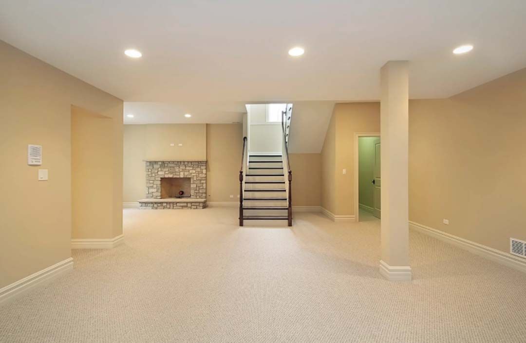 A beige carpeted basement with a fireplace, stairs, and a green door. Recessed lighting illuminates the neutral-toned space.