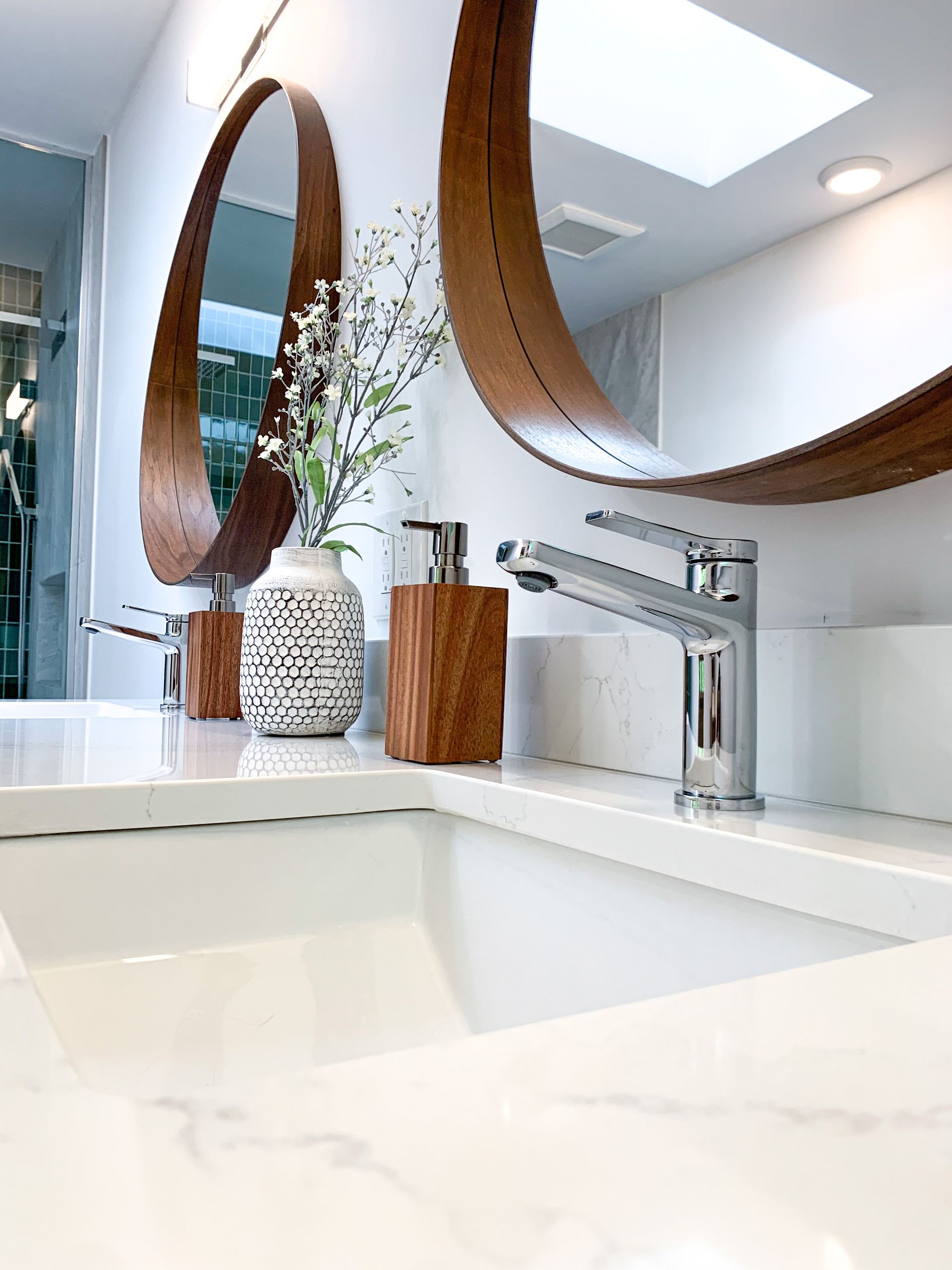 White bathroom with two round wood-framed mirrors, white countertops, and silver faucets. Decorative vase and wood soap dispenser.