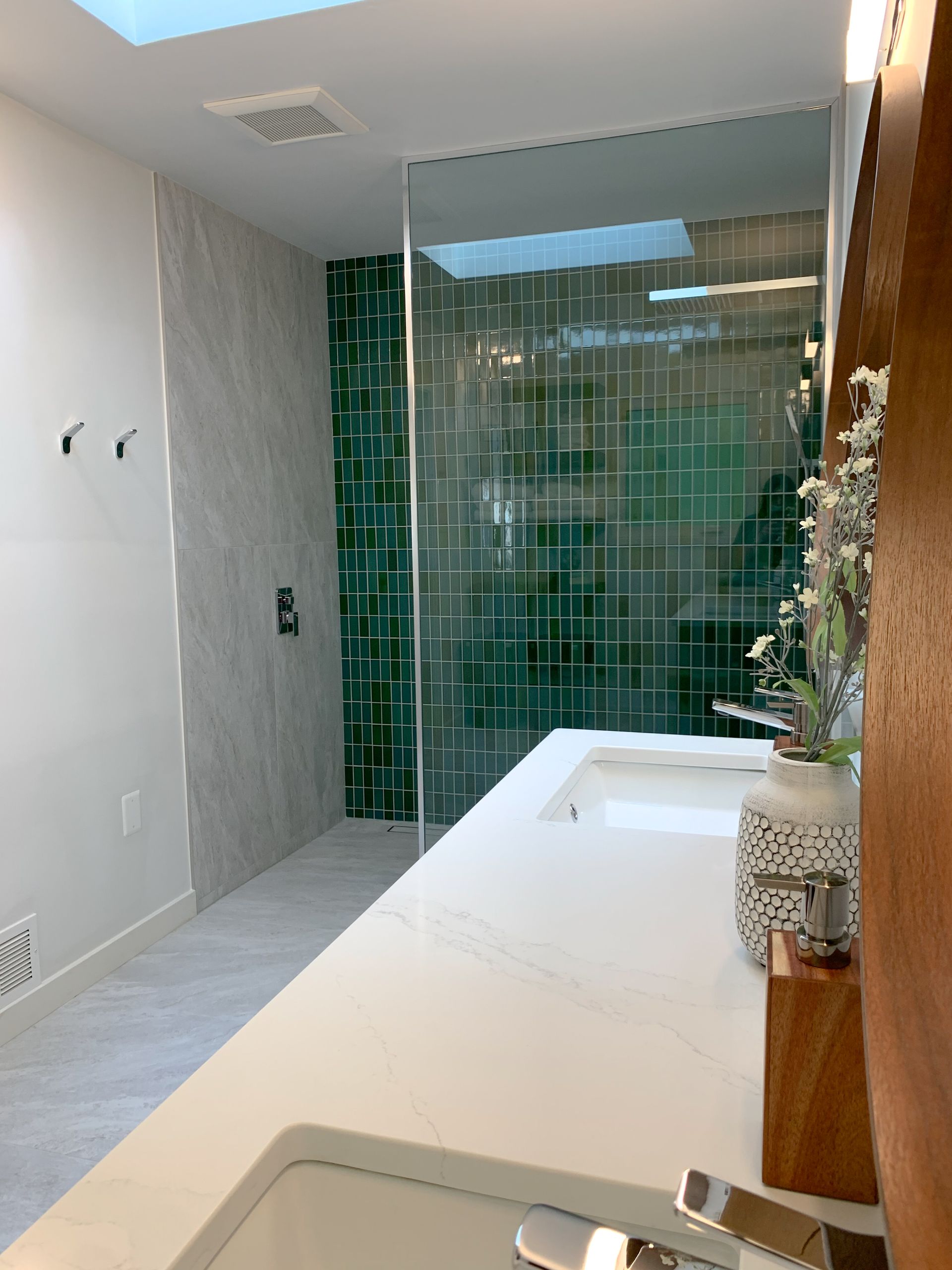 A modern bathroom with a white countertop and sink. A green tiled shower with a glass door is visible, alongside a gray tiled wall.