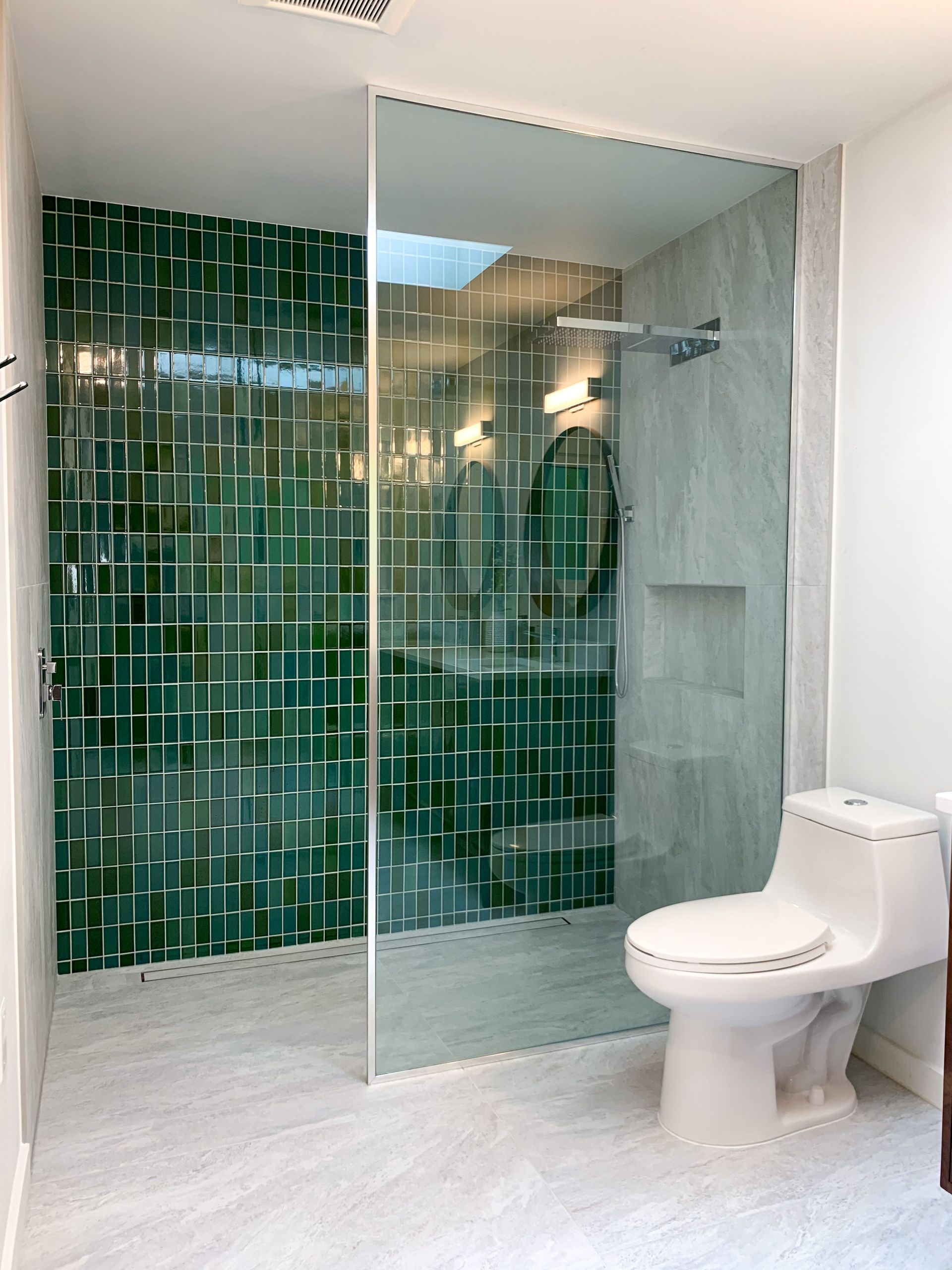 A modern bathroom with a green tile shower, glass door, and white toilet. The shower has a built-in niche and a silver showerhead.