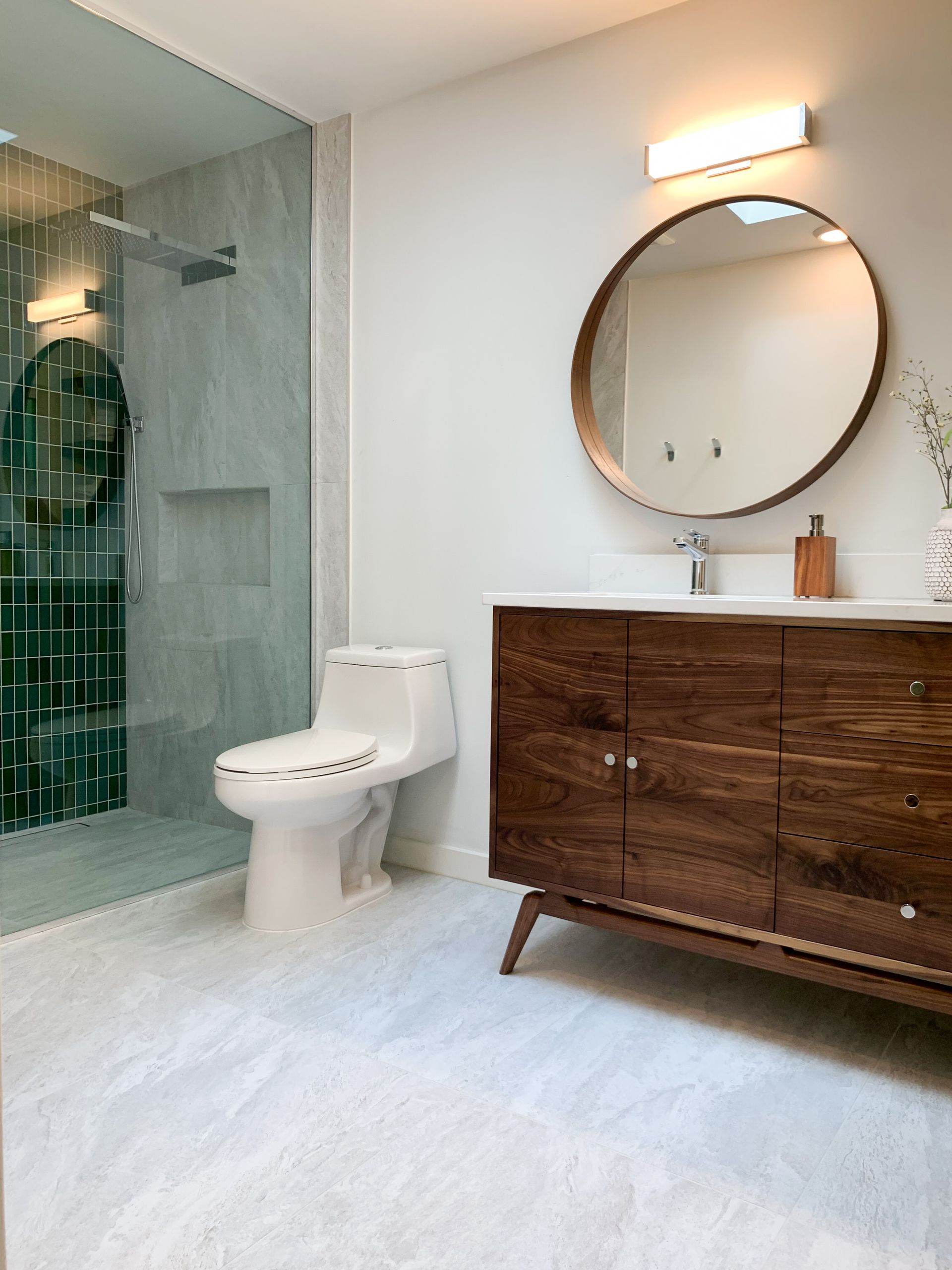 Modern bathroom with a wooden vanity, round mirror, and a walk-in shower with green tile accents.